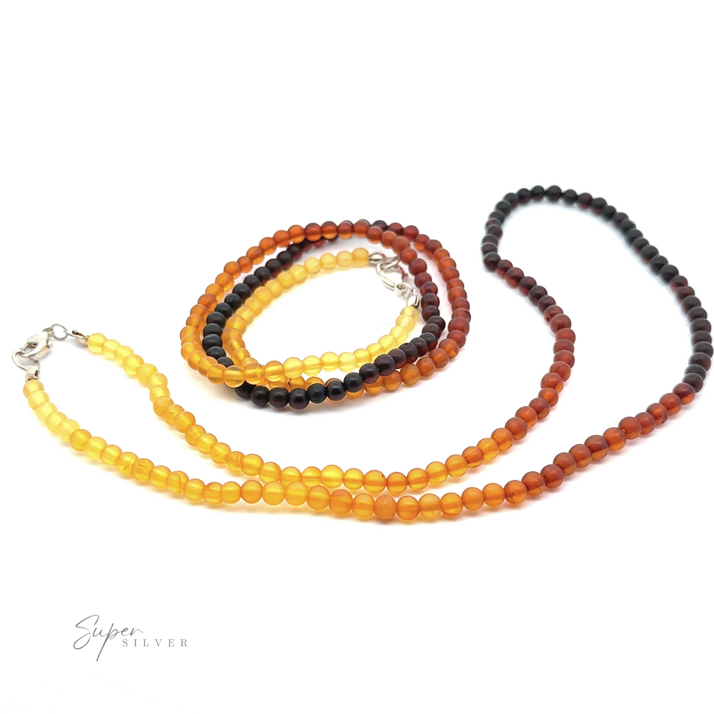 Three Ombre Beaded Amber Necklaces in varying shades of yellow to brown are displayed on a white background. The necklaces feature small round beads and sterling silver clasps, with one coiled in a circular shape.