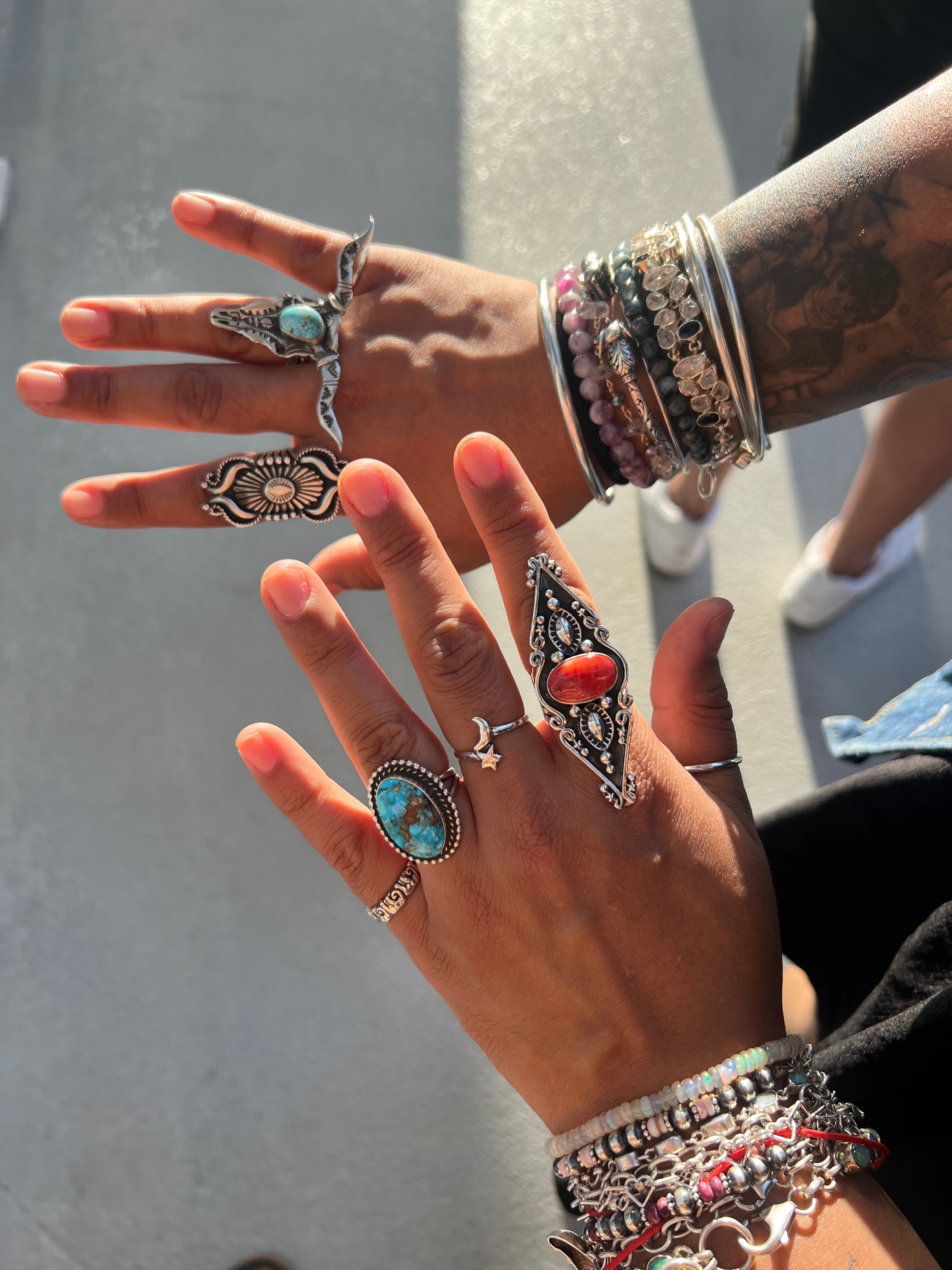 Two tattooed hands, adorned with bracelets and bangles, showcase the Stunning Native American Statement Ring as sunlight shines—a true tribute to Southwest artistry.
