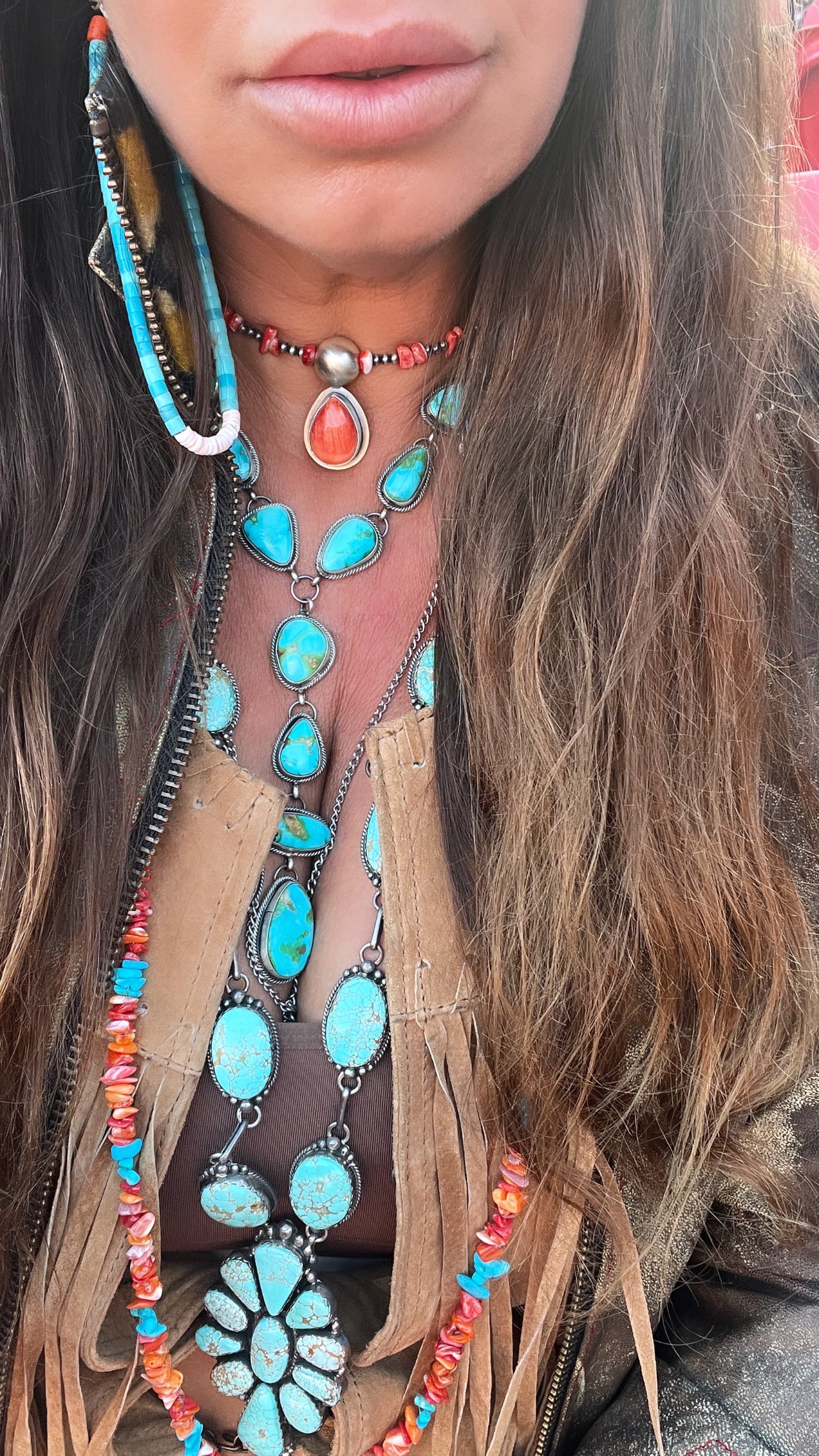 A person wearing multiple turquoise and coral stone jewelry pieces, a fringed top, a feather earring, and a Wrap Around Spiny Oyster Shell Choker Necklace crafted by Native American artisans.