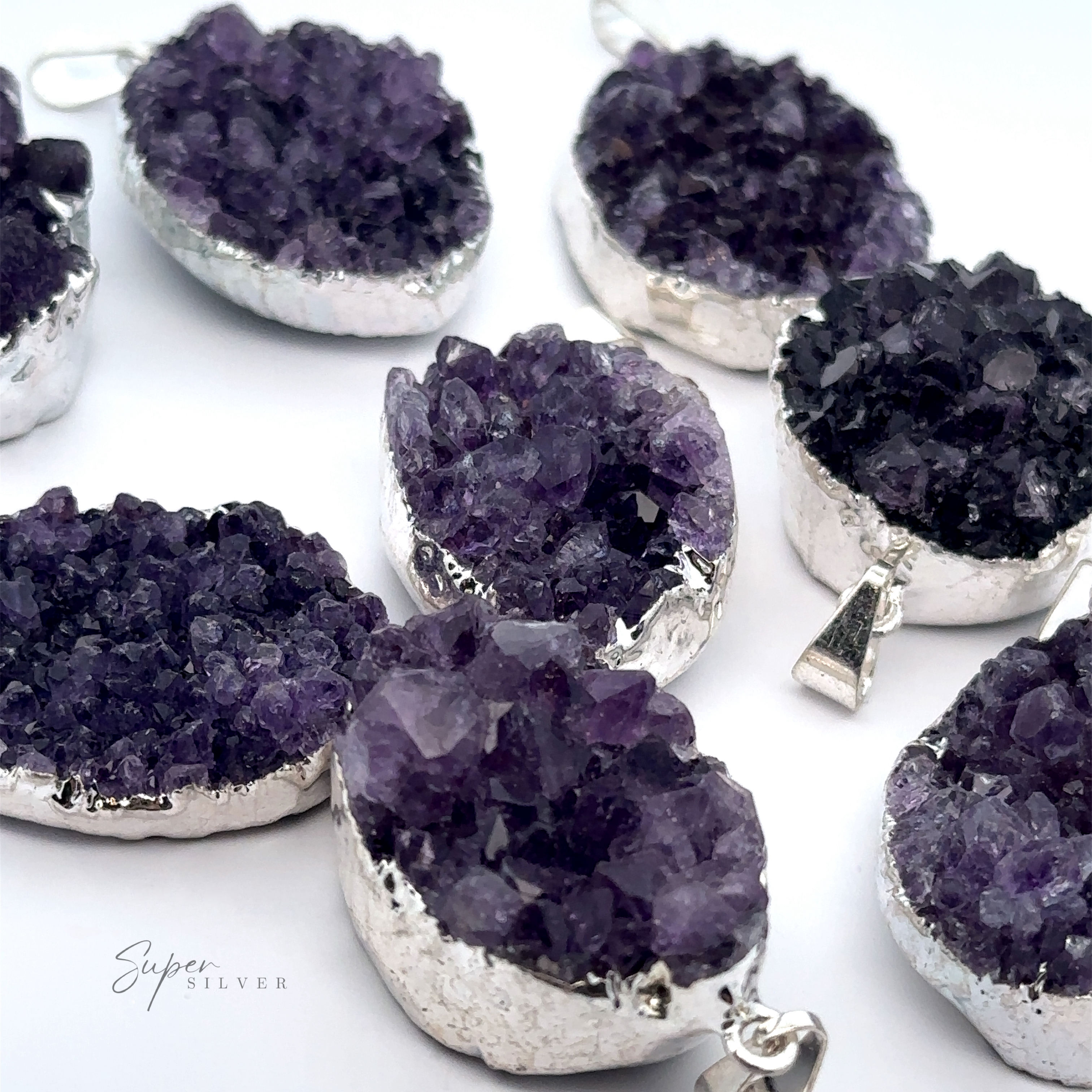 Amethyst Geode Pendants with silver plated settings are displayed on a white surface. Each pendant features a rough, organic texture and rich purple hues, making every piece unique.