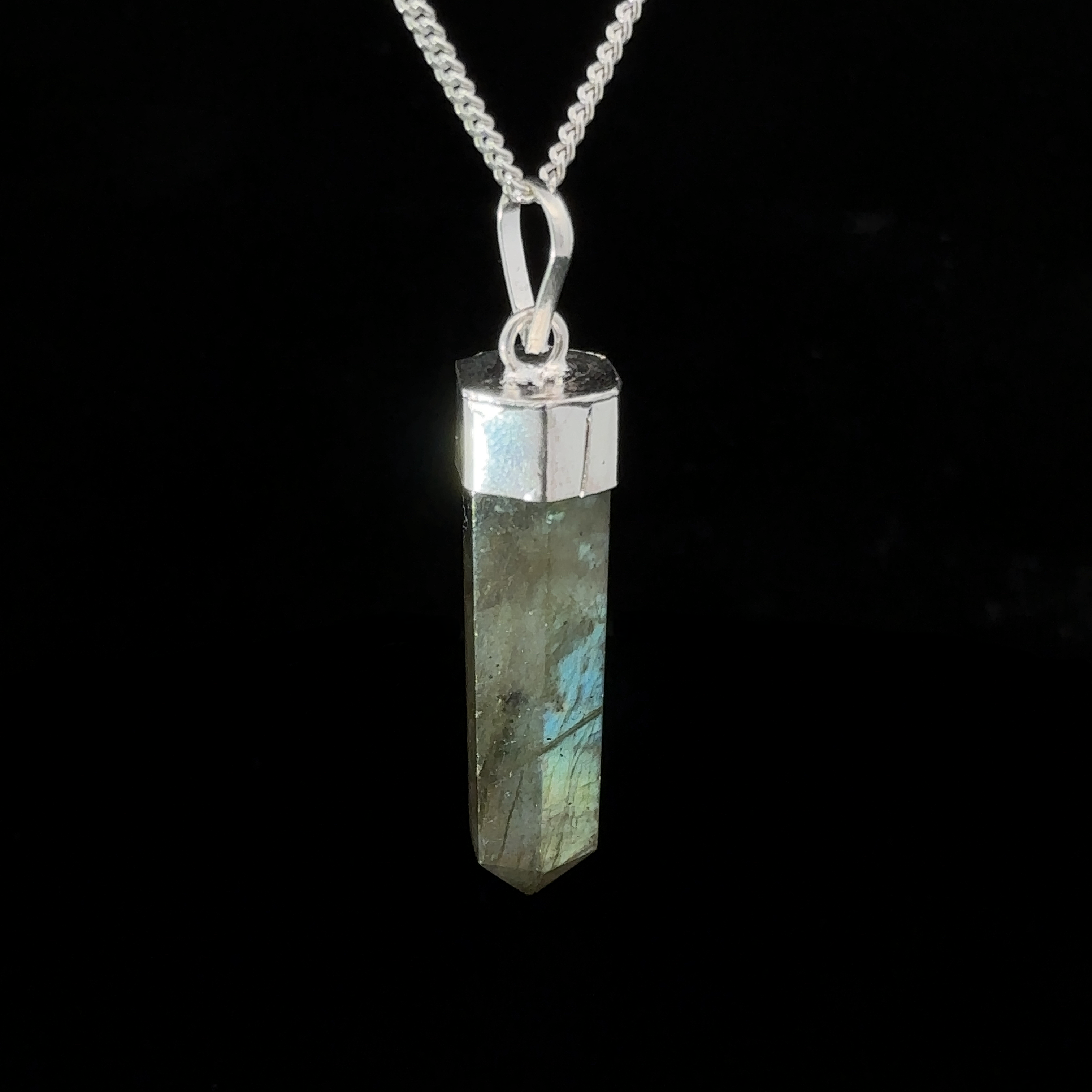 A Simple Gemstone Point Pendant featuring a long, hexagonal labradorite crystal with metallic hues, set in a silver-colored cap, hanging on a silver chain. This boho vibe crystal pendant emanates mystical charm, perfect for any free spirit.