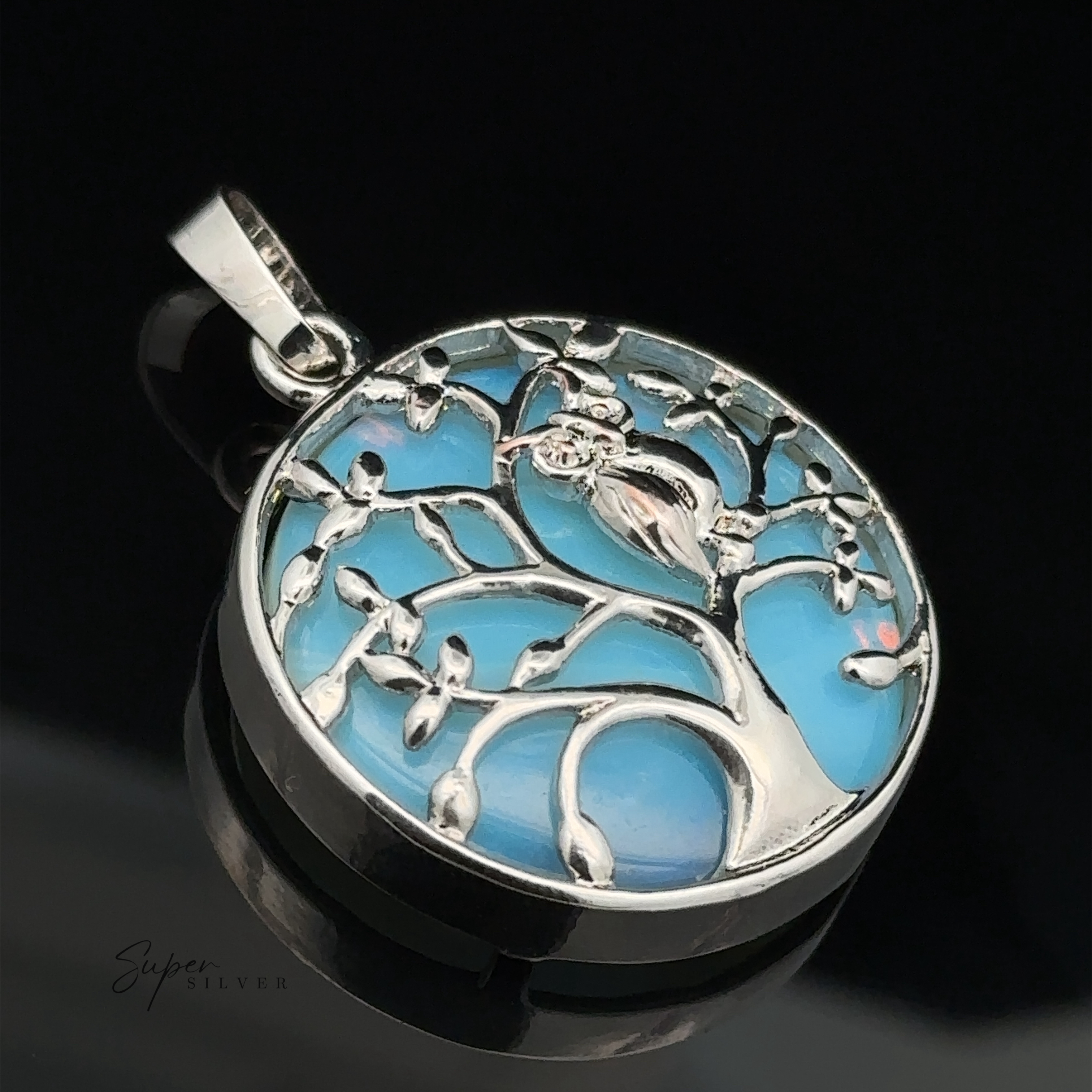 A round silver and mixed metals Owl and Tree Pendant featuring a bird perched on a branch within a stone tree design, set against a blue background.