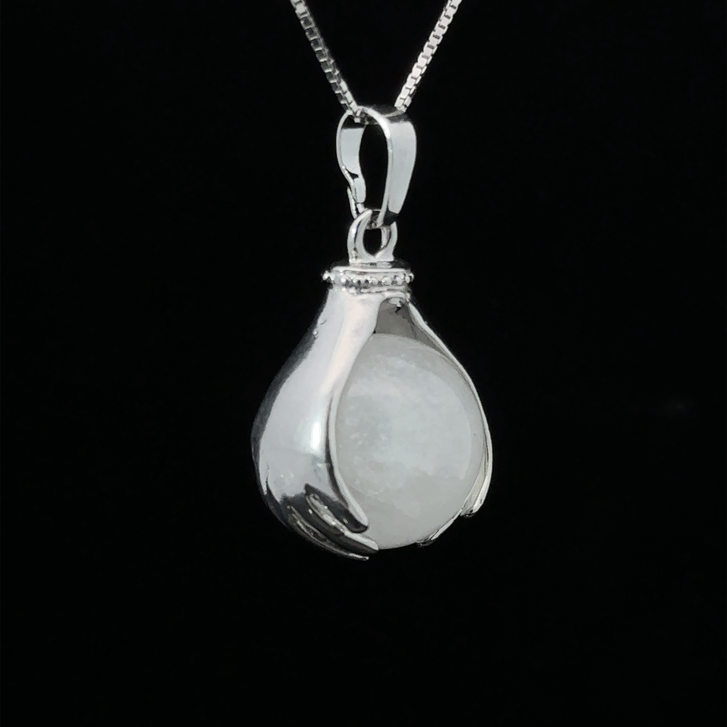 A Sphere Crystal Pendant features two hands delicately holding a round quartz stone. The pendant hangs from a fine silver chain against a black background.