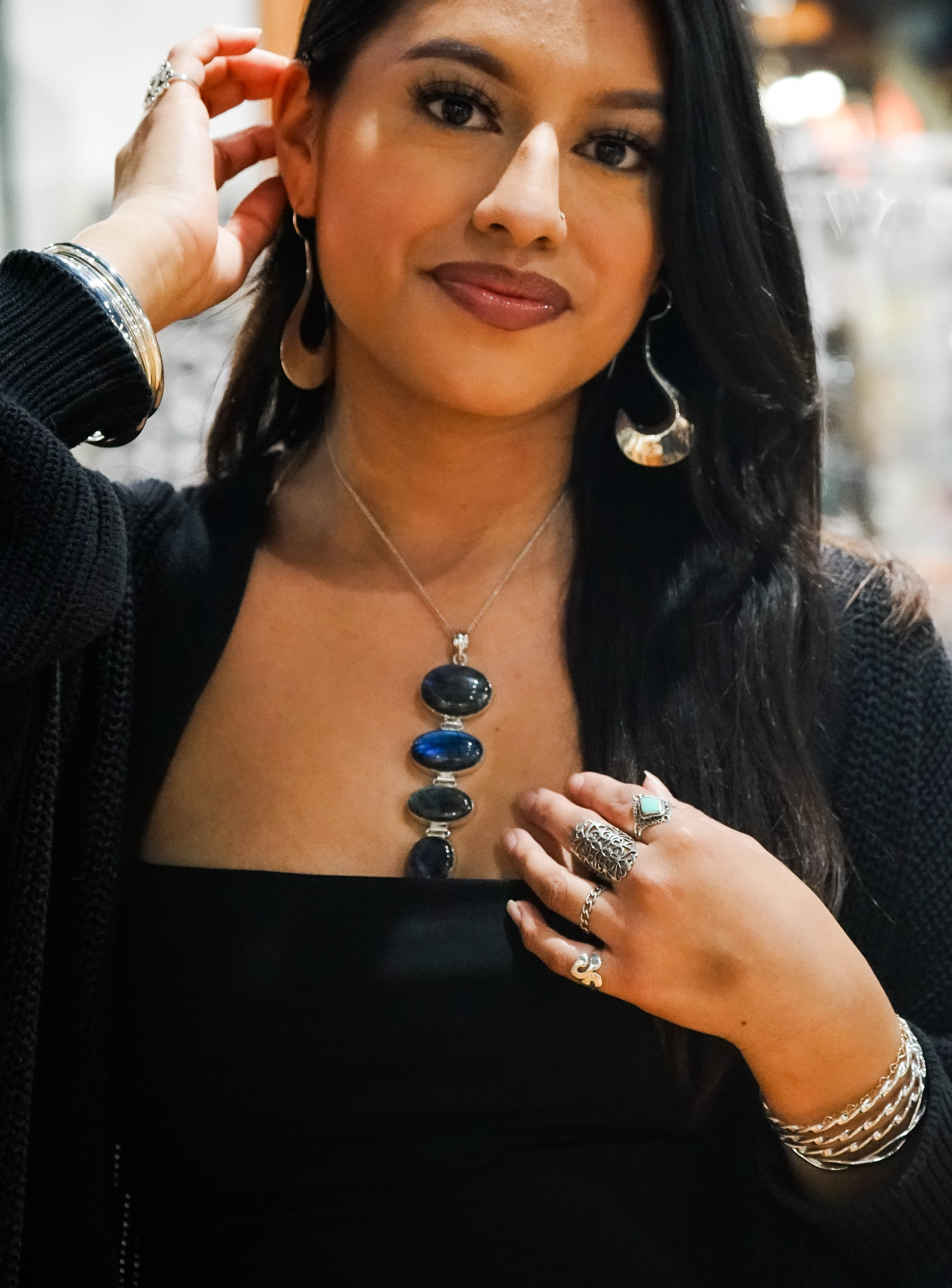 A woman wearing large hoop earrings, multiple rings, and a Tiered Statement Labradorite Pendant adjusts her earring while looking at the camera.