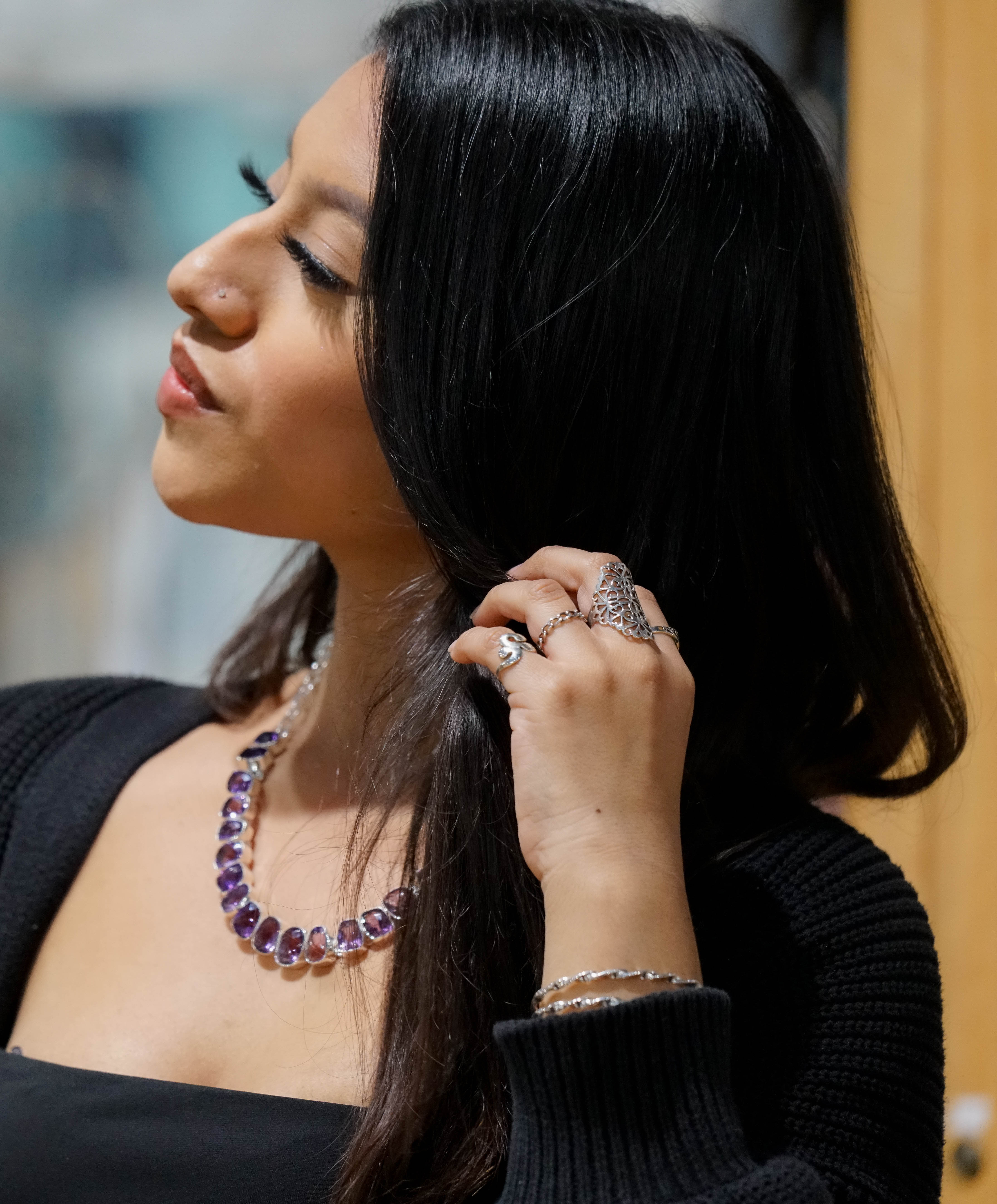 A woman with long dark hair, wearing a black top and a Statement Gemstone Necklace adorned with amethyst beads, rings, and a nose stud, holds a strand of her hair while looking to the side.