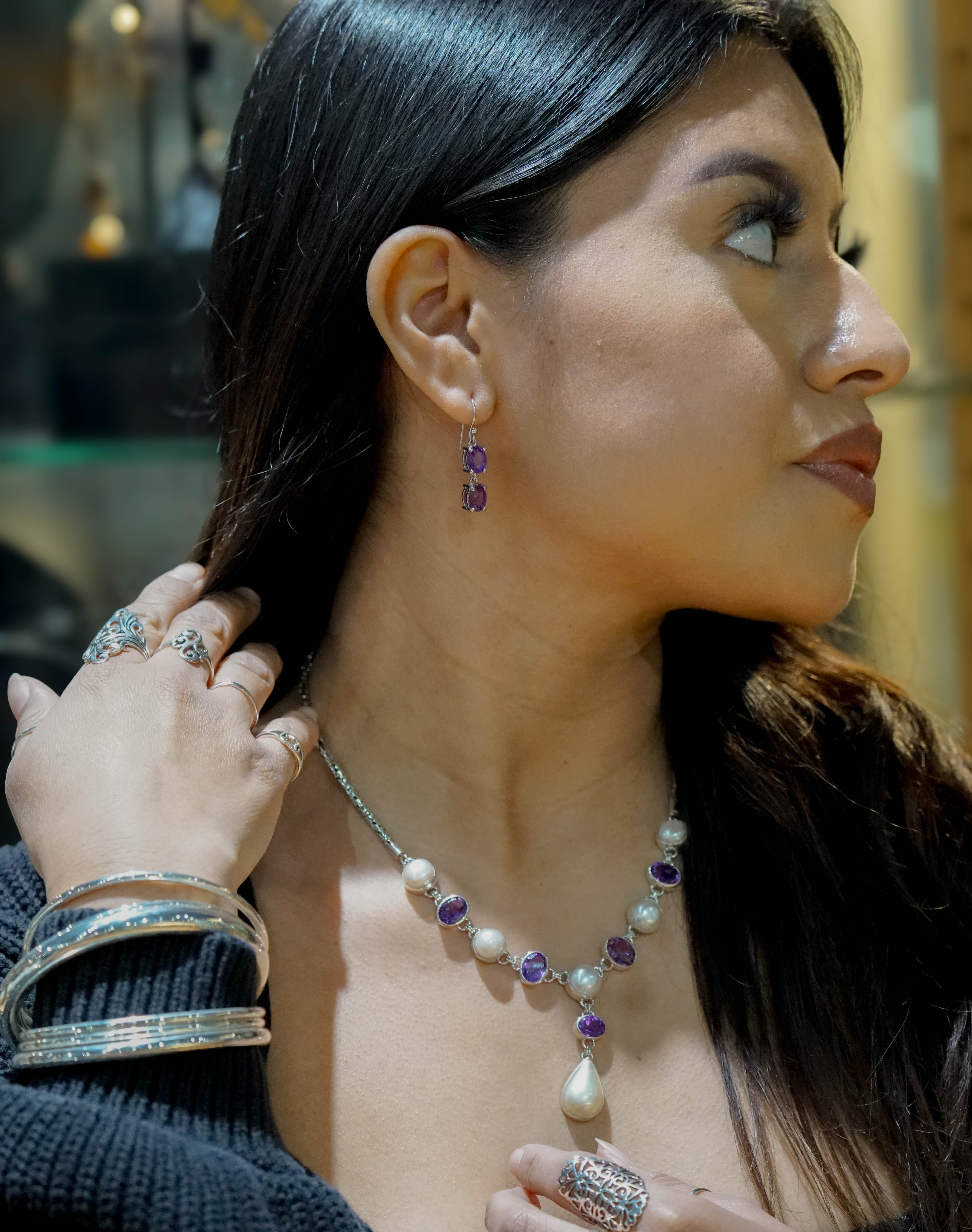A woman with long dark hair is wearing a Stunning Pearl and Gemstone Statement Necklace, matching earrings, and multiple bracelets and rings. She is looking to the right and touching her hair with her left hand.