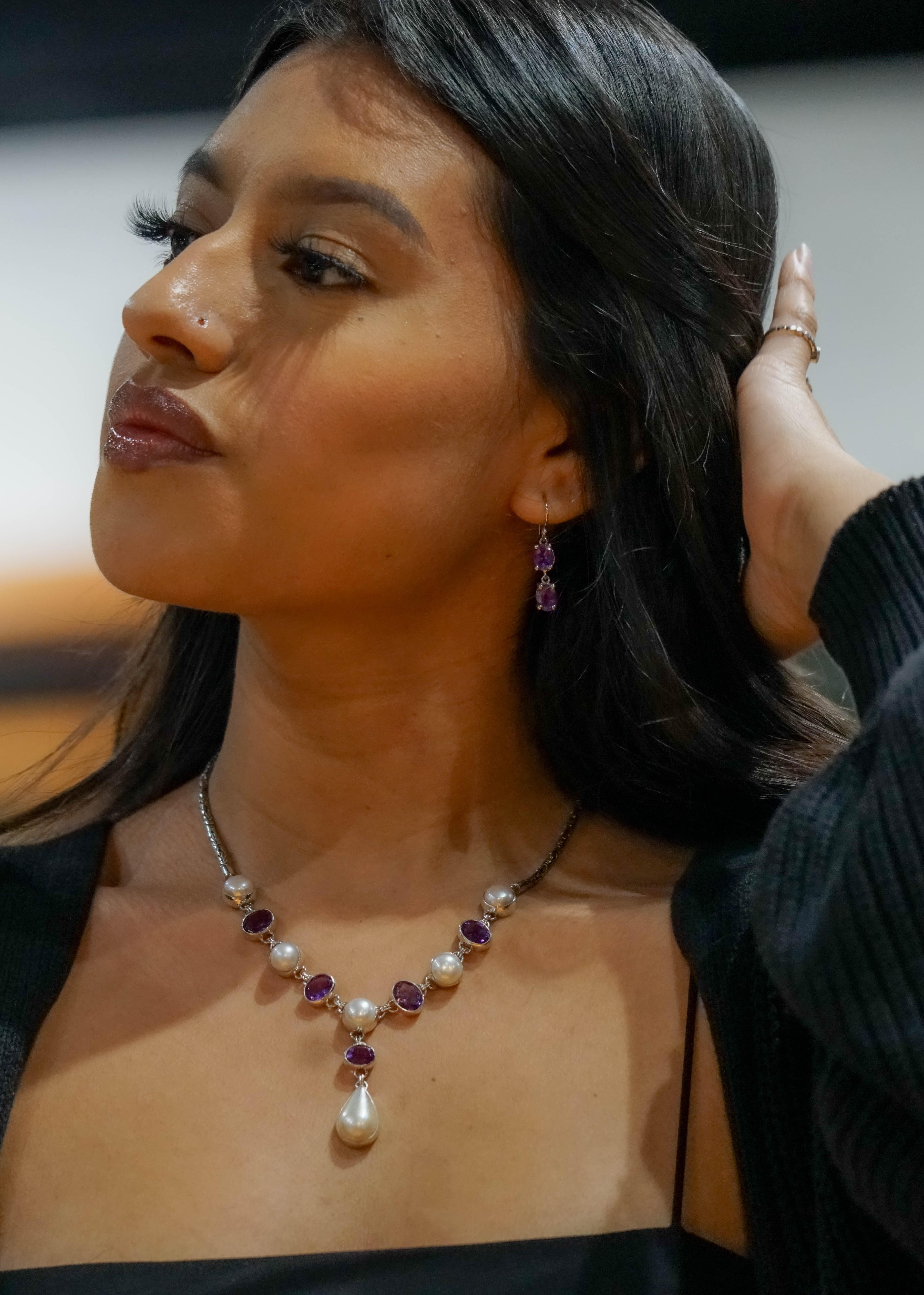 A woman wearing a black top displays a stunning Pearl and Gemstone Statement Necklace, along with matching earrings. She has long, dark hair and appears to be adjusting it with one hand.