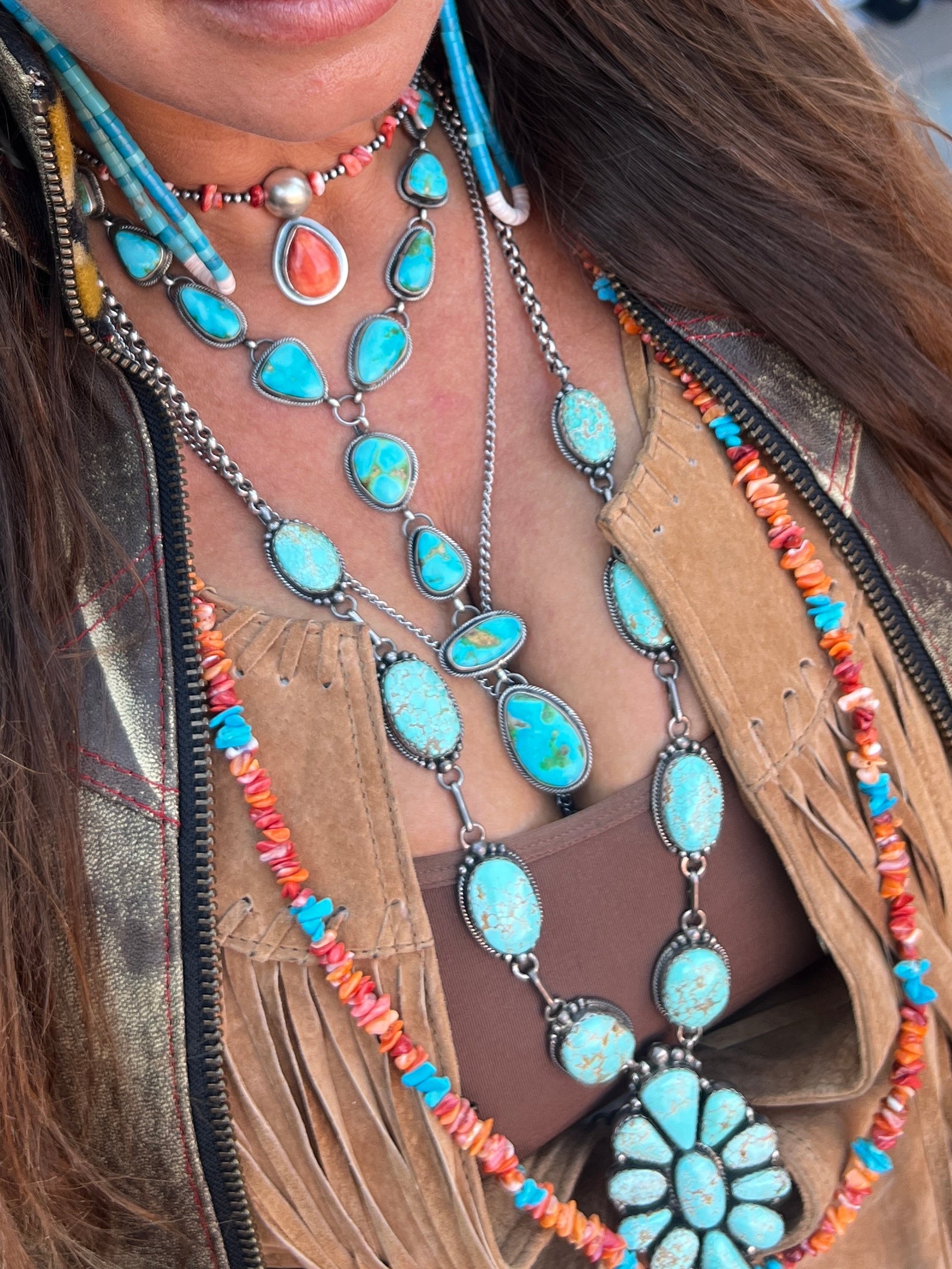 Close-up of a person wearing the Stunning Sonora Gold Lariat Necklace And Earrings Set adorned with authentic Native American turquoise stones, coral beads, and silver details, paired with a brown fringed suede jacket.