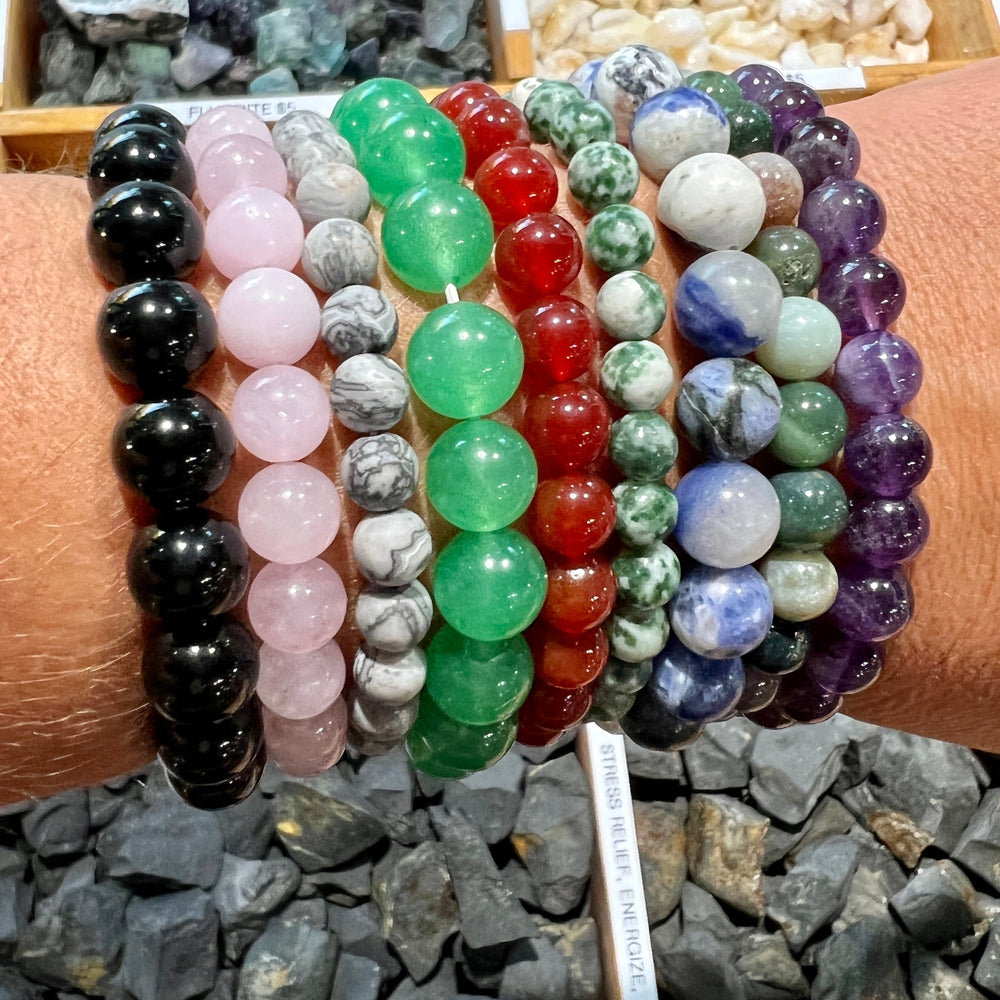 
                  
                    A person displaying multiple colorful 4mm Beaded Stone Bracelets on their wrist, above a selection of scattered stones.
                  
                