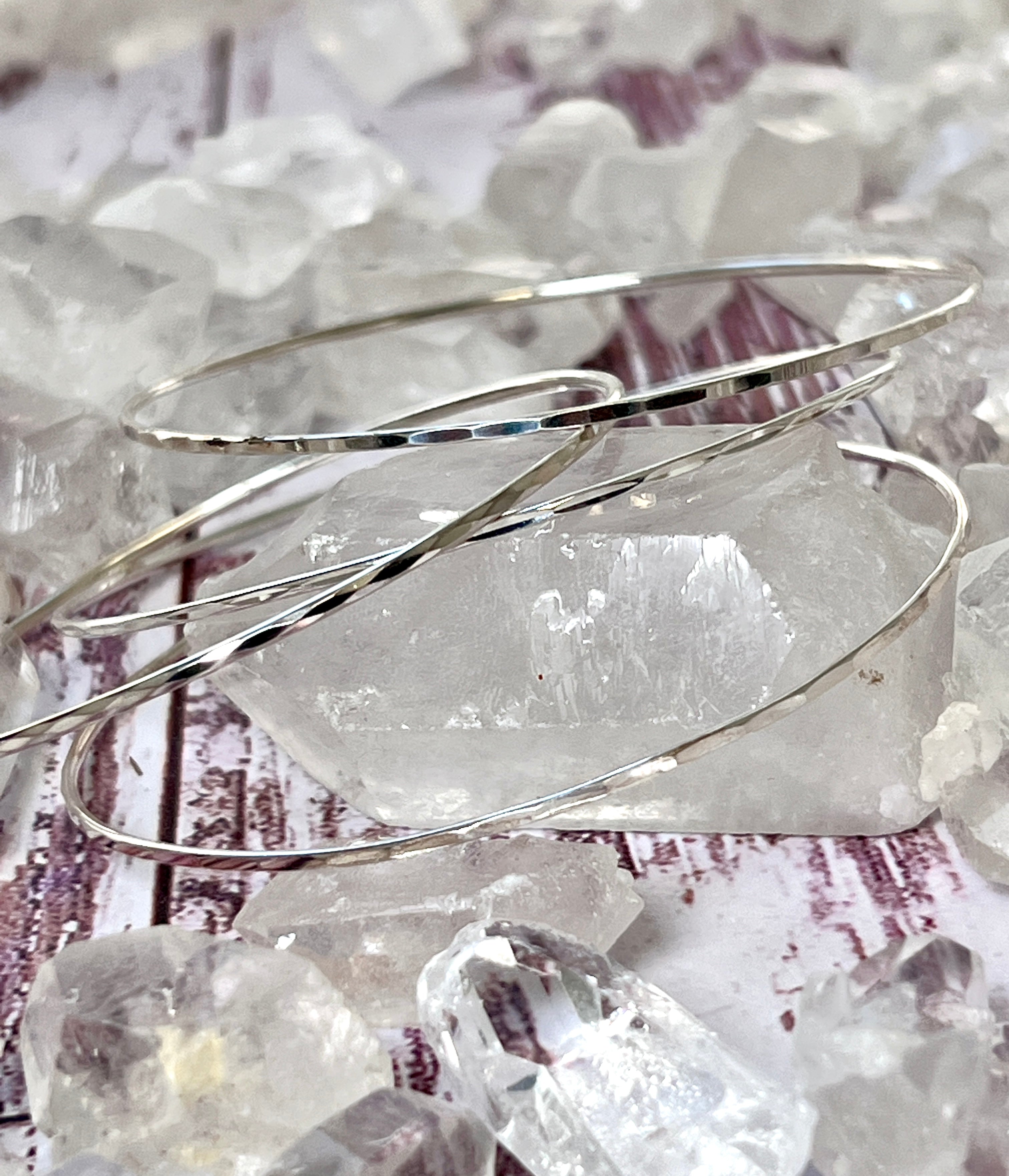 Three Faceted Silver Bangle Bracelets rest on a cluster of translucent quartz crystals on a wooden surface.