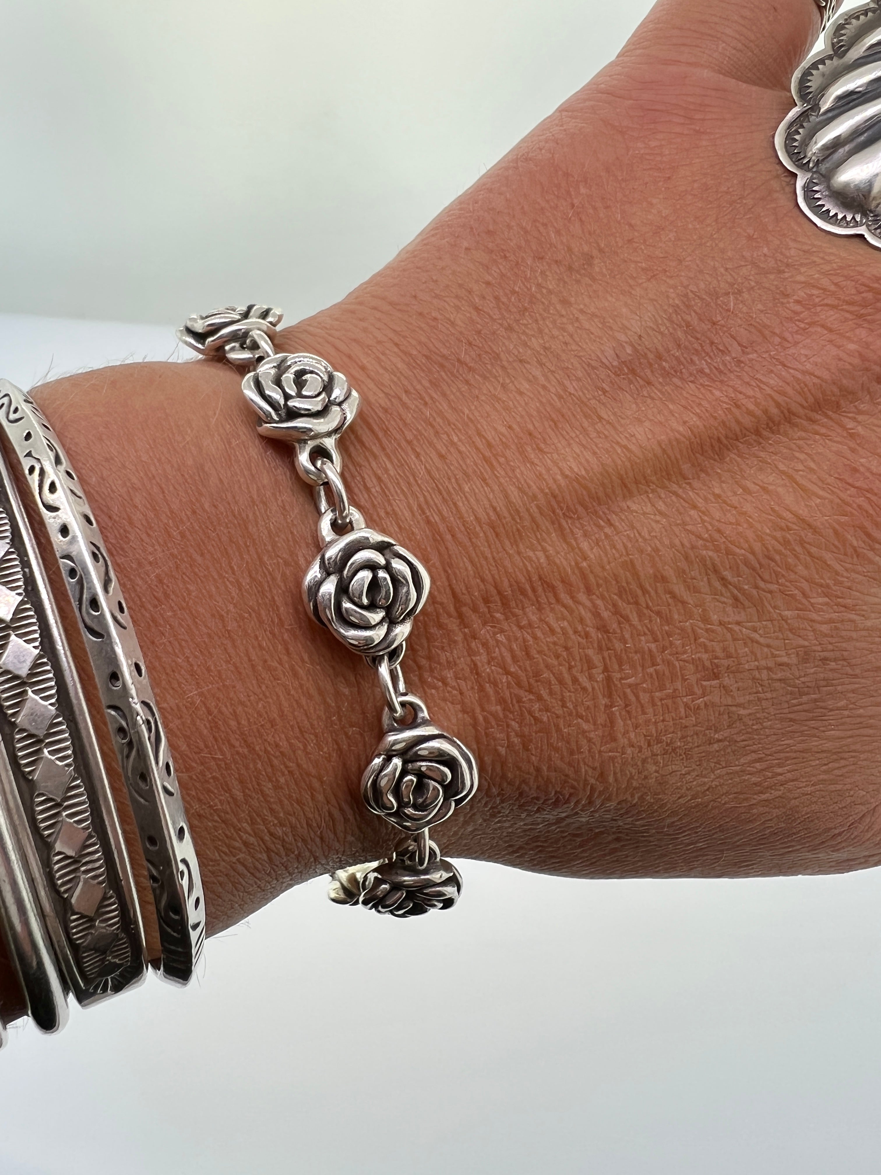 A wrist adorned with three silver bracelets, including a Chic Link Rose Bracelet with vintage vibes and another with intricate patterns. The thumb is partially visible wearing a large, detailed .925 sterling silver ring.