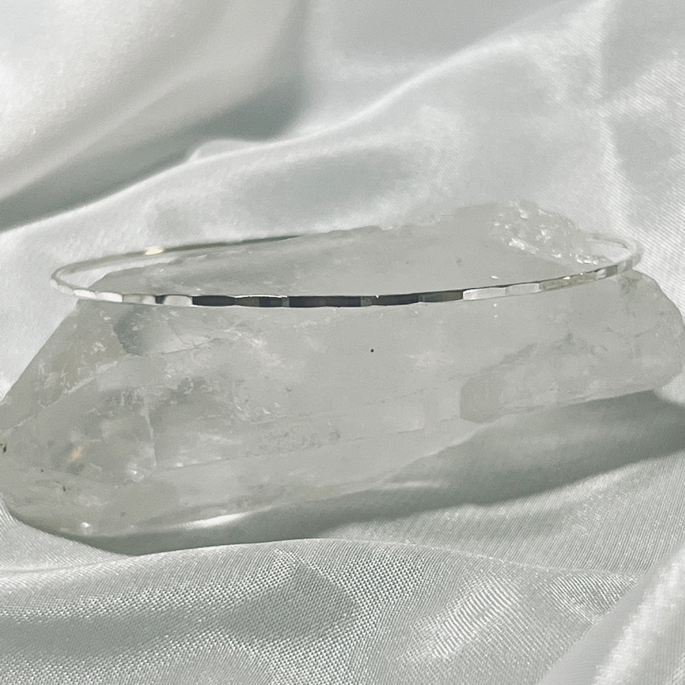 
                  
                    A thin Faceted Silver Bangle Bracelet rests on a large, faceted cut clear quartz crystal, positioned on a white satin fabric background.
                  
                