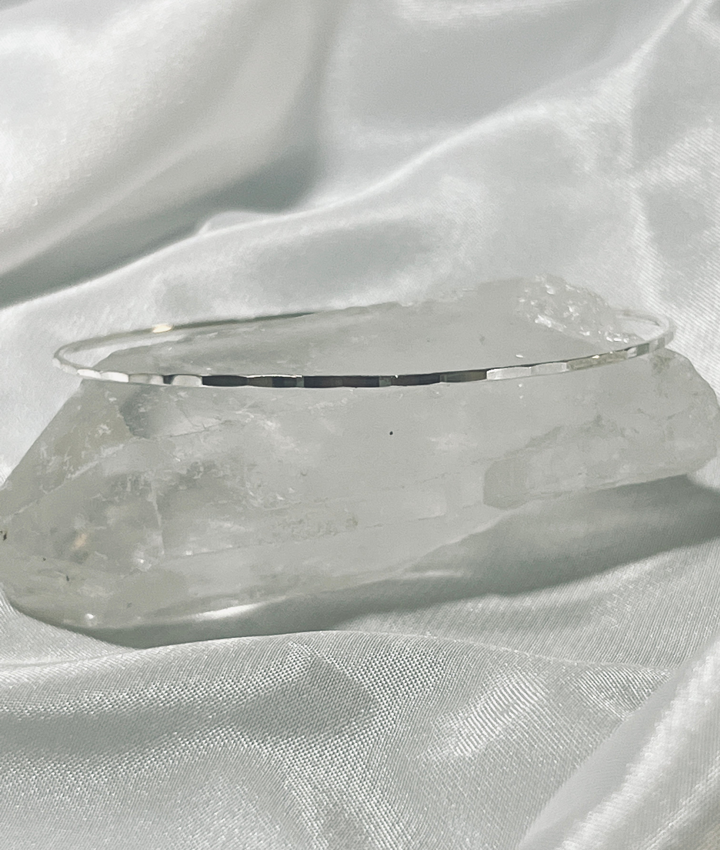 A thin Faceted Silver Bangle Bracelet rests on a large, faceted cut clear quartz crystal, positioned on a white satin fabric background.