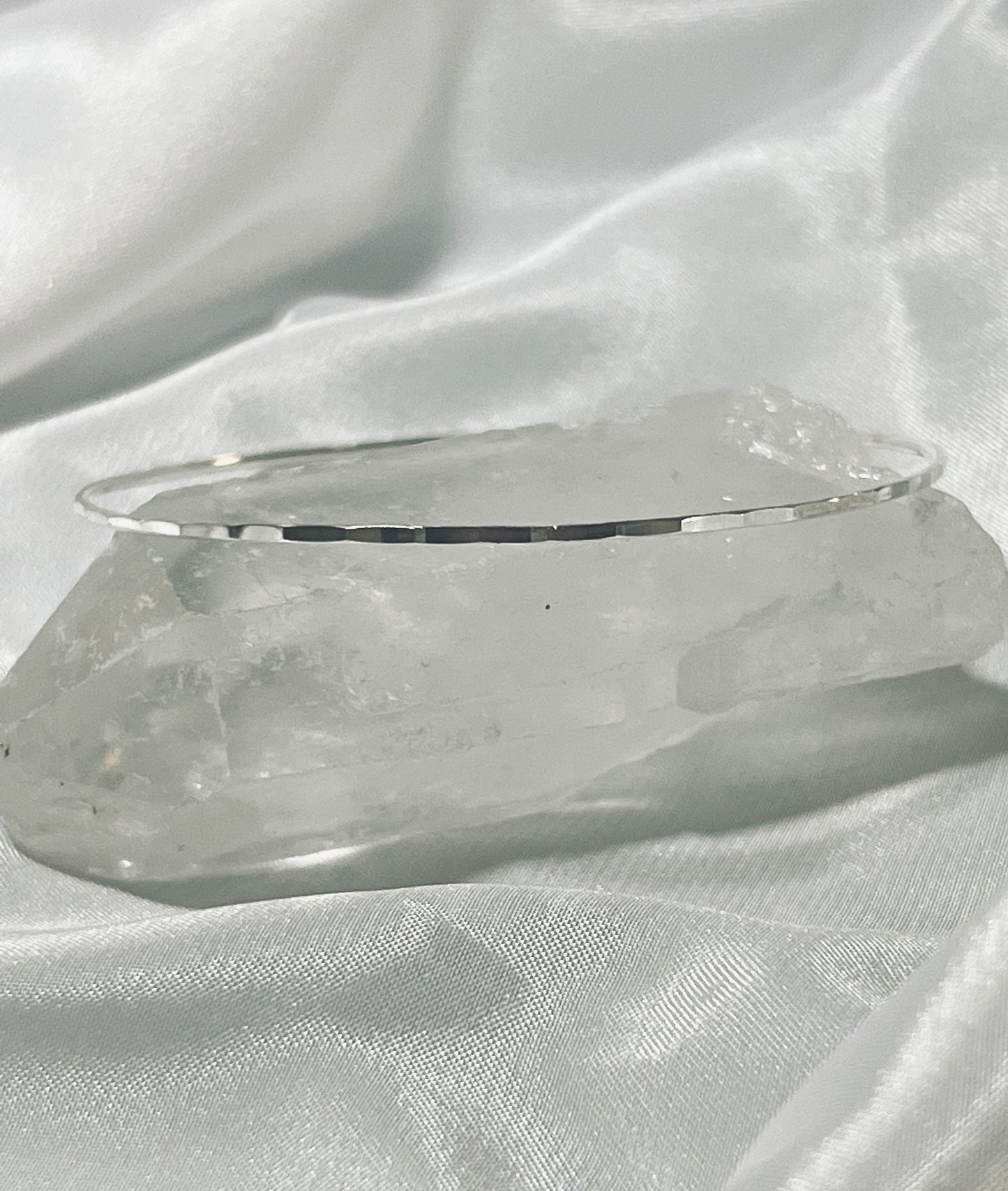 
                  
                    A thin Faceted Silver Bangle Bracelet rests on a large, faceted cut clear quartz crystal, positioned on a white satin fabric background.
                  
                