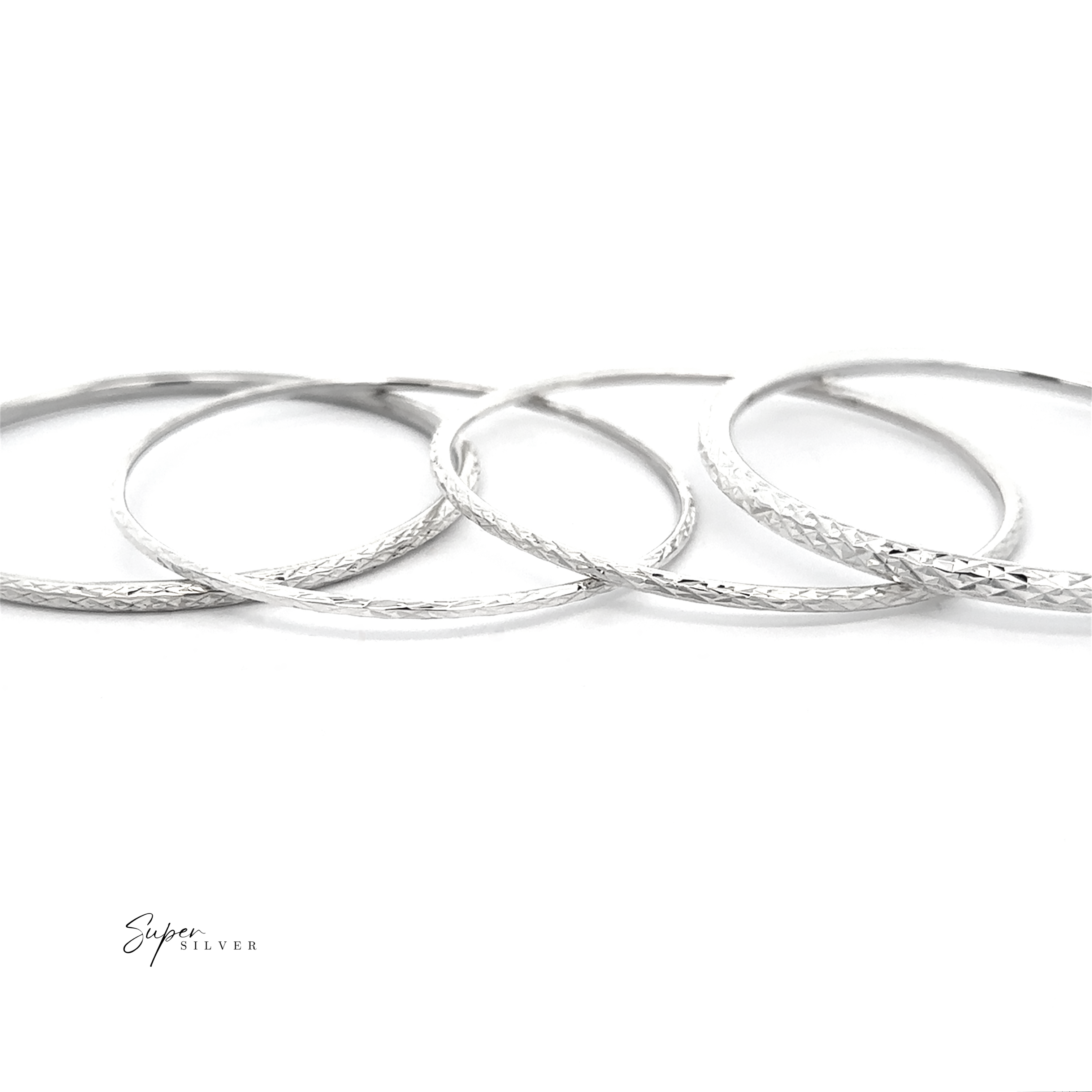 Five thin, textured bangles from The Spark Stack Bangles collection are artfully overlapped on a white background, shimmering in the light. "Super Silver" is displayed in the lower left corner.