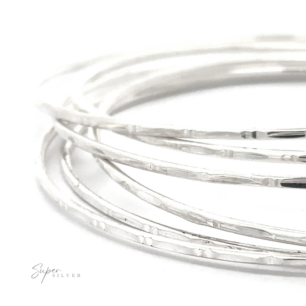 Close-up of several thin Delicate Faceted Cut Bangle Bracelets stacked together on a white surface. The texture appears slightly hammered, adding to their unique charm. "Super Silver" is written in small text in the lower left corner.