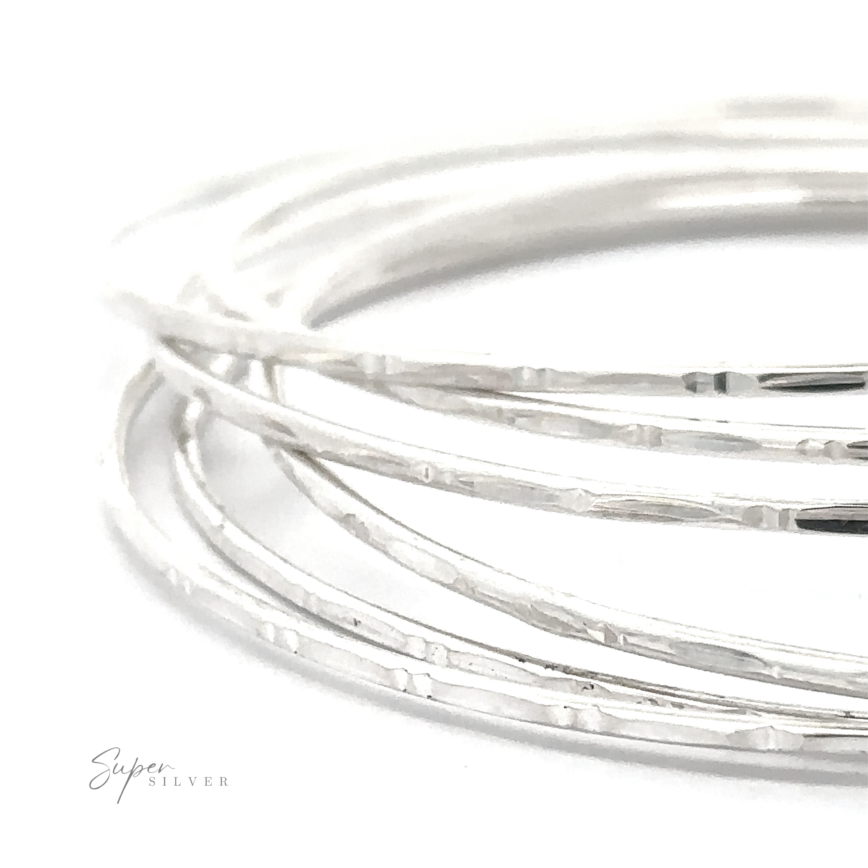 Close-up of several thin Delicate Faceted Cut Bangle Bracelets stacked together on a white surface. The texture appears slightly hammered, adding to their unique charm. "Super Silver" is written in small text in the lower left corner.