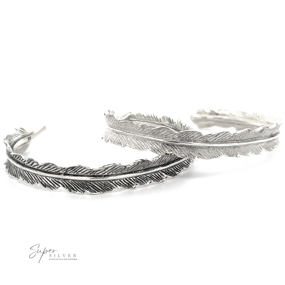 Two Detailed Feather Cuffs, one in dark silver and the other in light silver .925 sterling silver, are displayed side by side on a white background. The brand name 