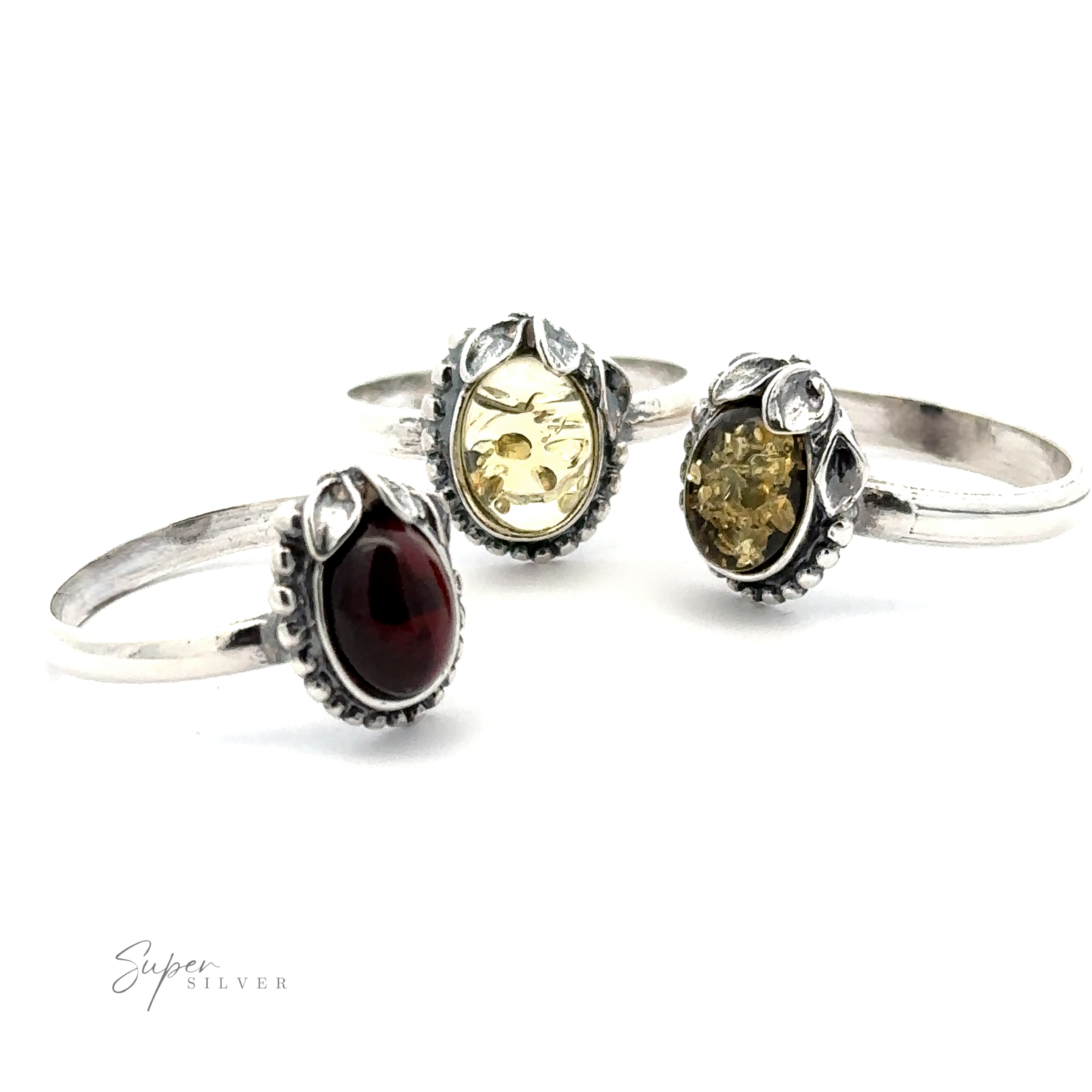 Three intricately designed silver rings with stone settings, including a red gemstone and two with yellow-hued stones, exude vintage elegance. Displayed on a white background, the "Super Silver" logo is visible in the lower left corner. One ring stands out as an Amber Ring with Beaded Border and Peace Lily Details, epitomizing nature-inspired jewelry.