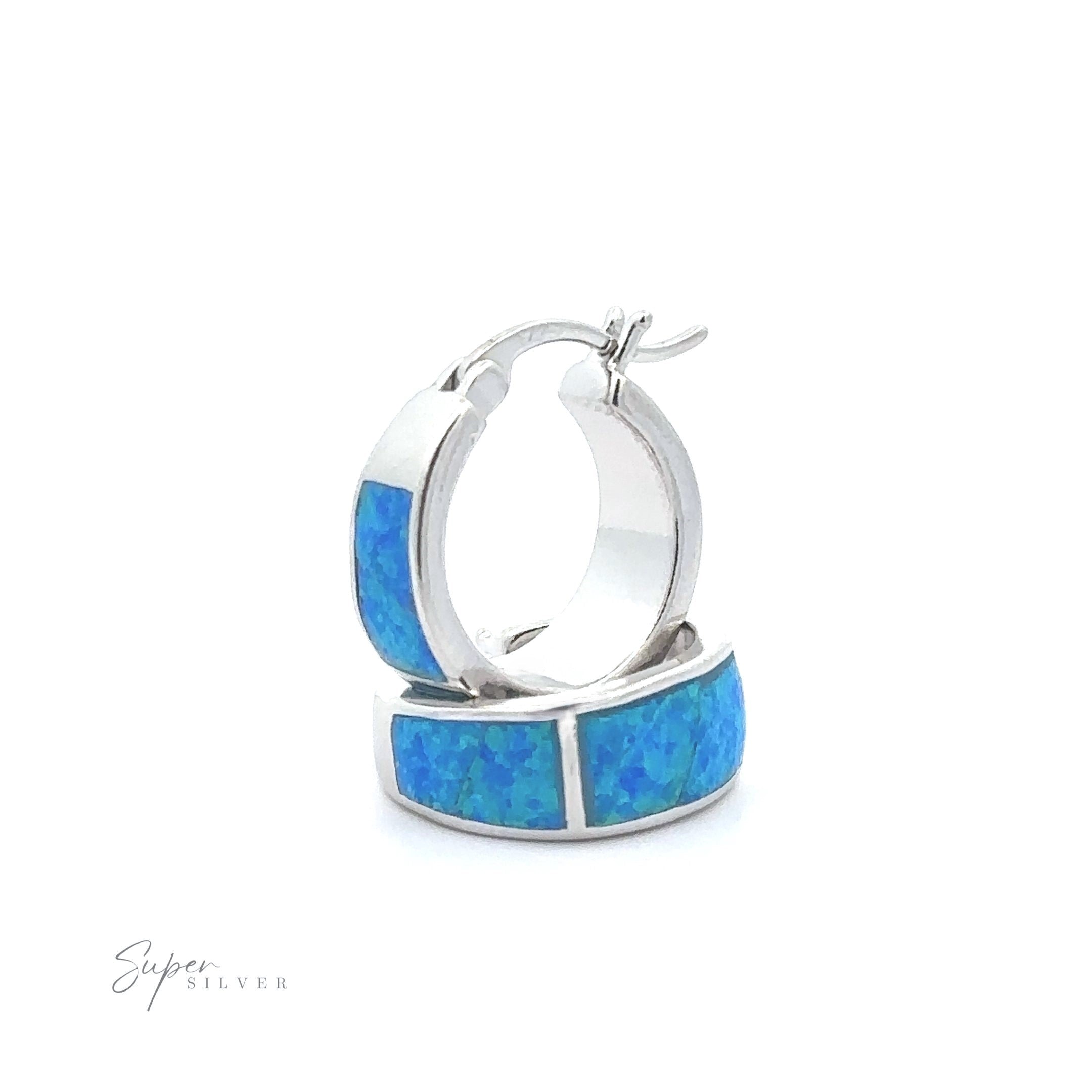 A pair of Blue Lab-Created Opal Huggie Hoop Earrings with silver inlays on a white background. The earrings, showcasing a stunning rhodium finish, are displayed standing upright and slightly overlapping. The "Super Silver" logo is visible in the corner.