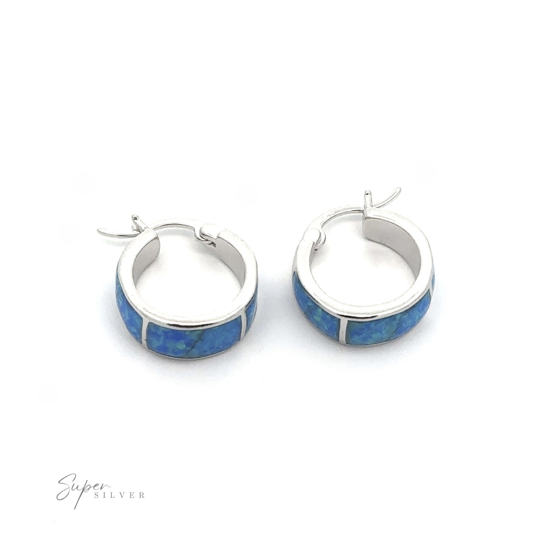 A pair of Blue Lab-Created Opal Huggie Hoop Earrings with blue lab-created opals set against a white background. The rhodium finish adds extra shine, and the logo "Super Silver" is visible at the bottom left.