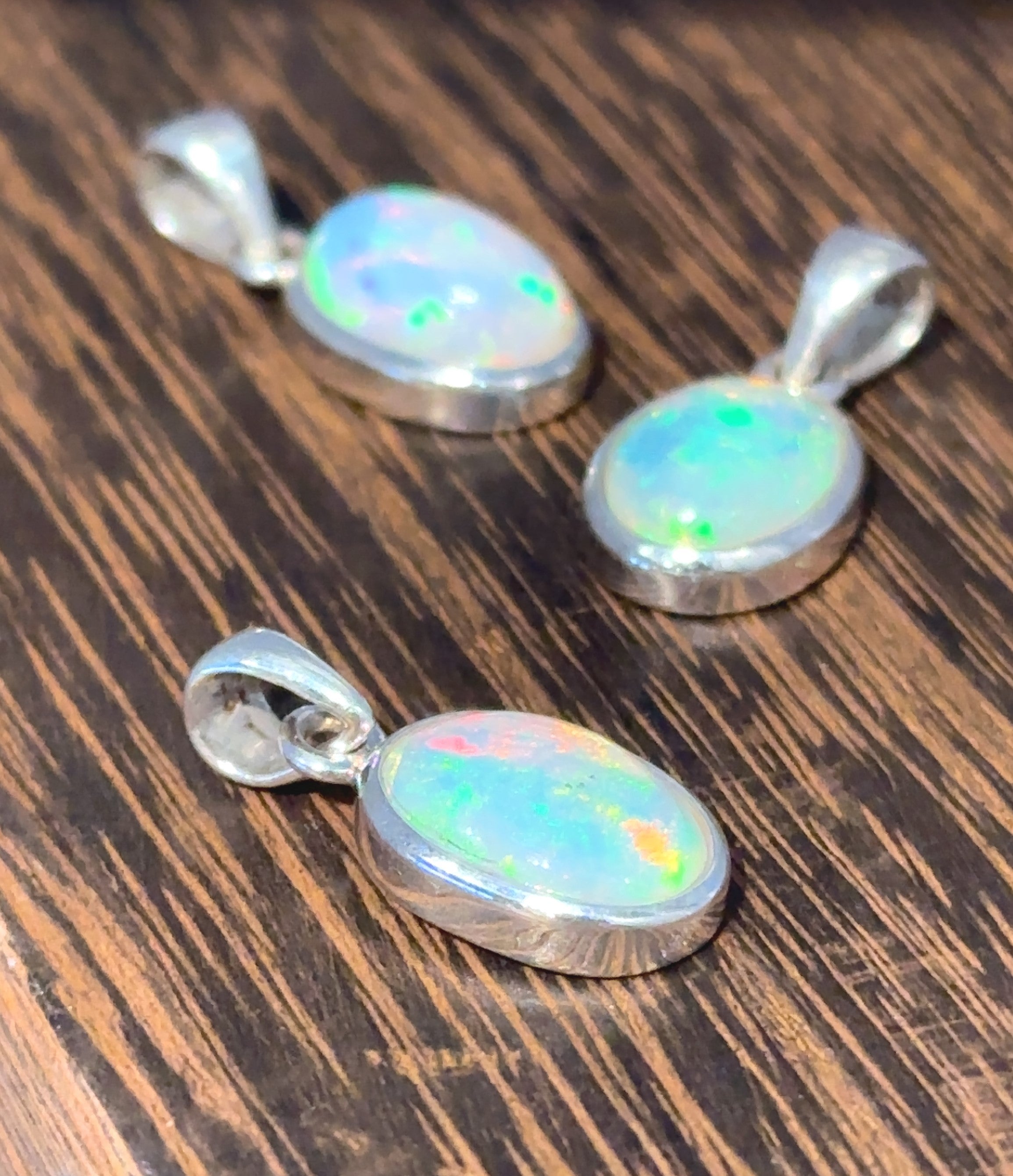 Three Oval Ethiopian Opal Pendants in sterling silver settings are elegantly displayed on a wooden surface, embodying a minimalist style.