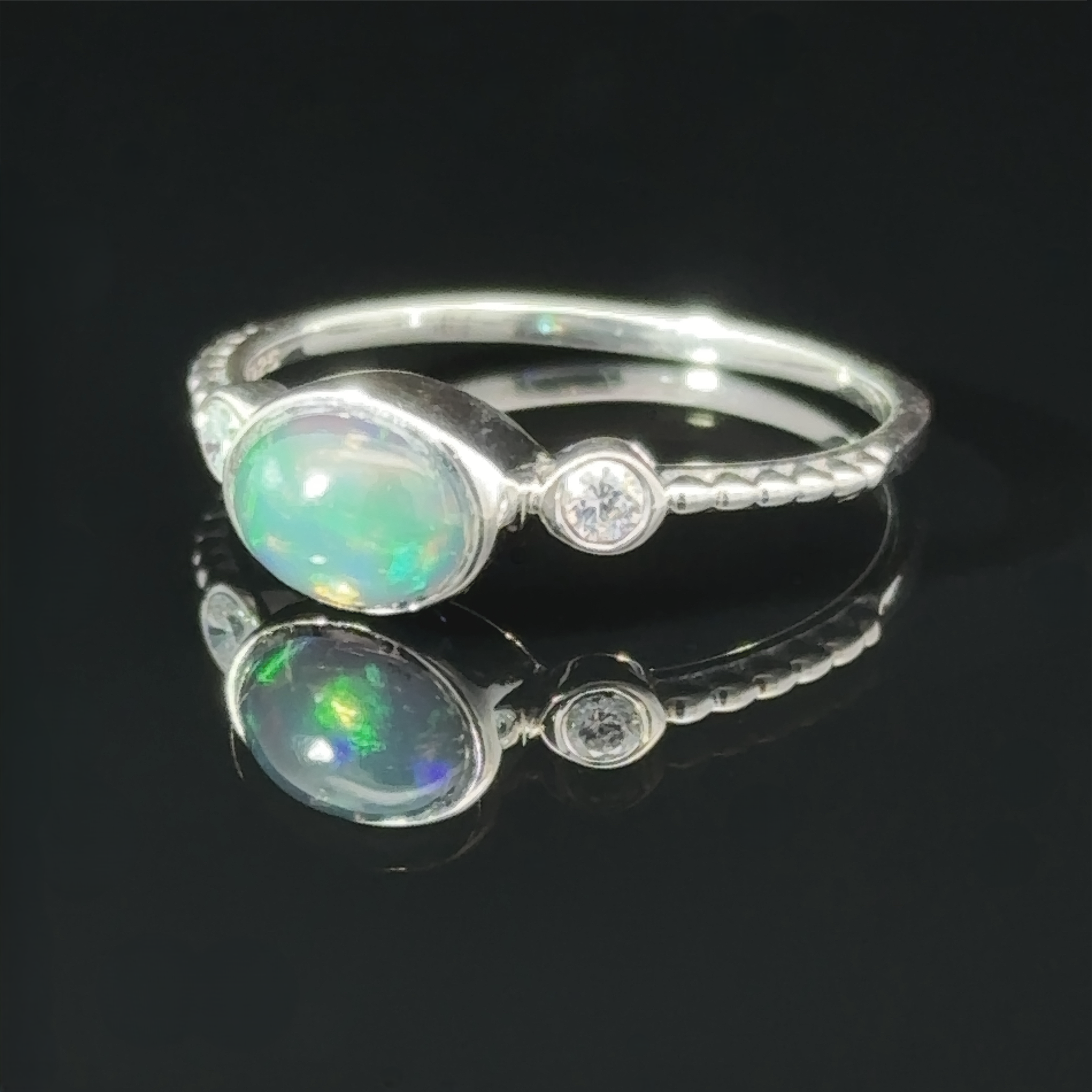Horizontal Oval Gemstone Ring with Beaded Band with an oval opal set next to a small round diamond, displayed on a reflective black surface.