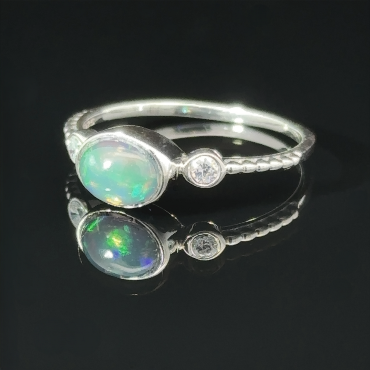 
                  
                    Horizontal Oval Gemstone Ring with Beaded Band with an oval opal set next to a small round diamond, displayed on a reflective black surface.
                  
                