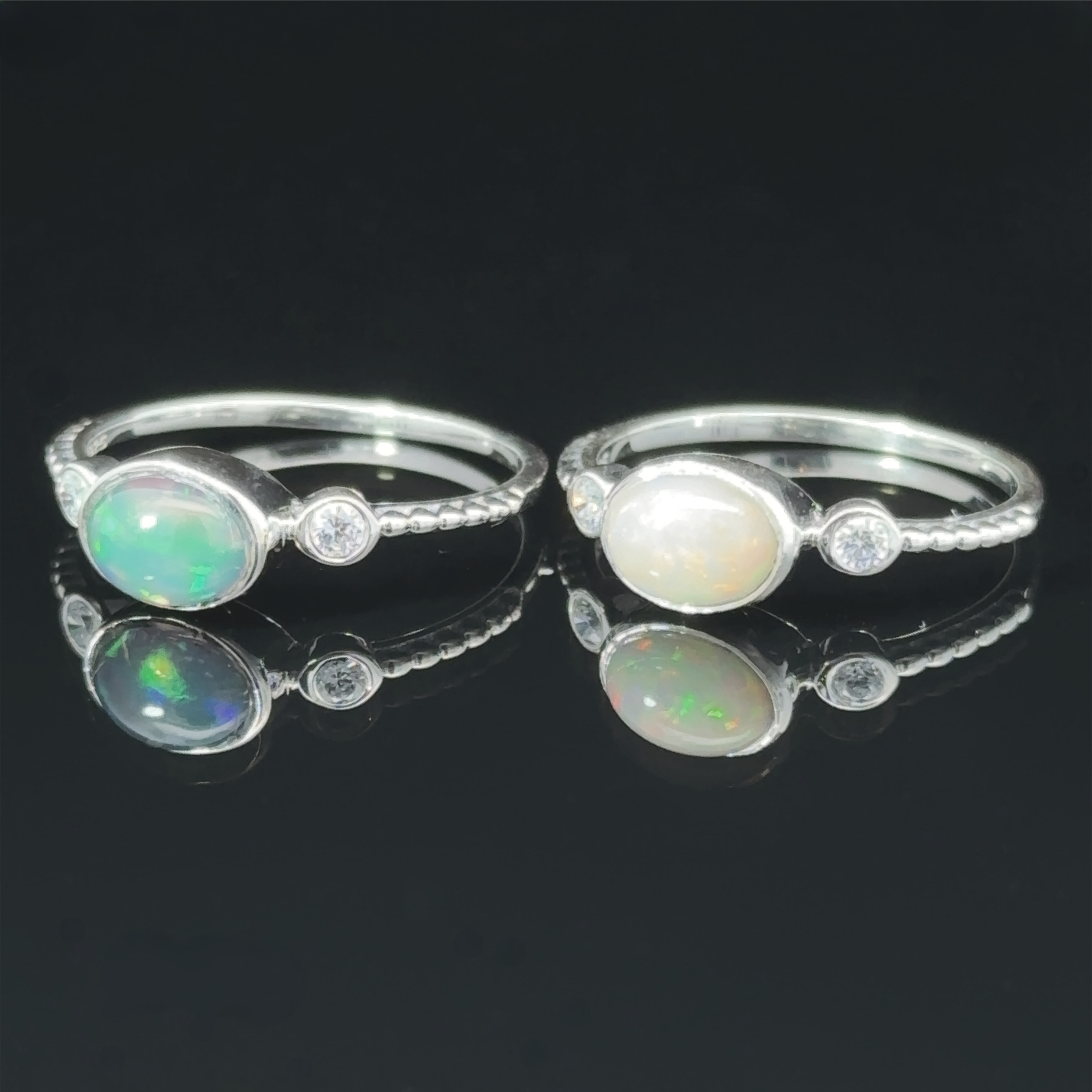 Two handcrafted Horizontal Oval Gemstone Rings with small diamond accents on a reflective black surface, showcasing the stones' iridescence.