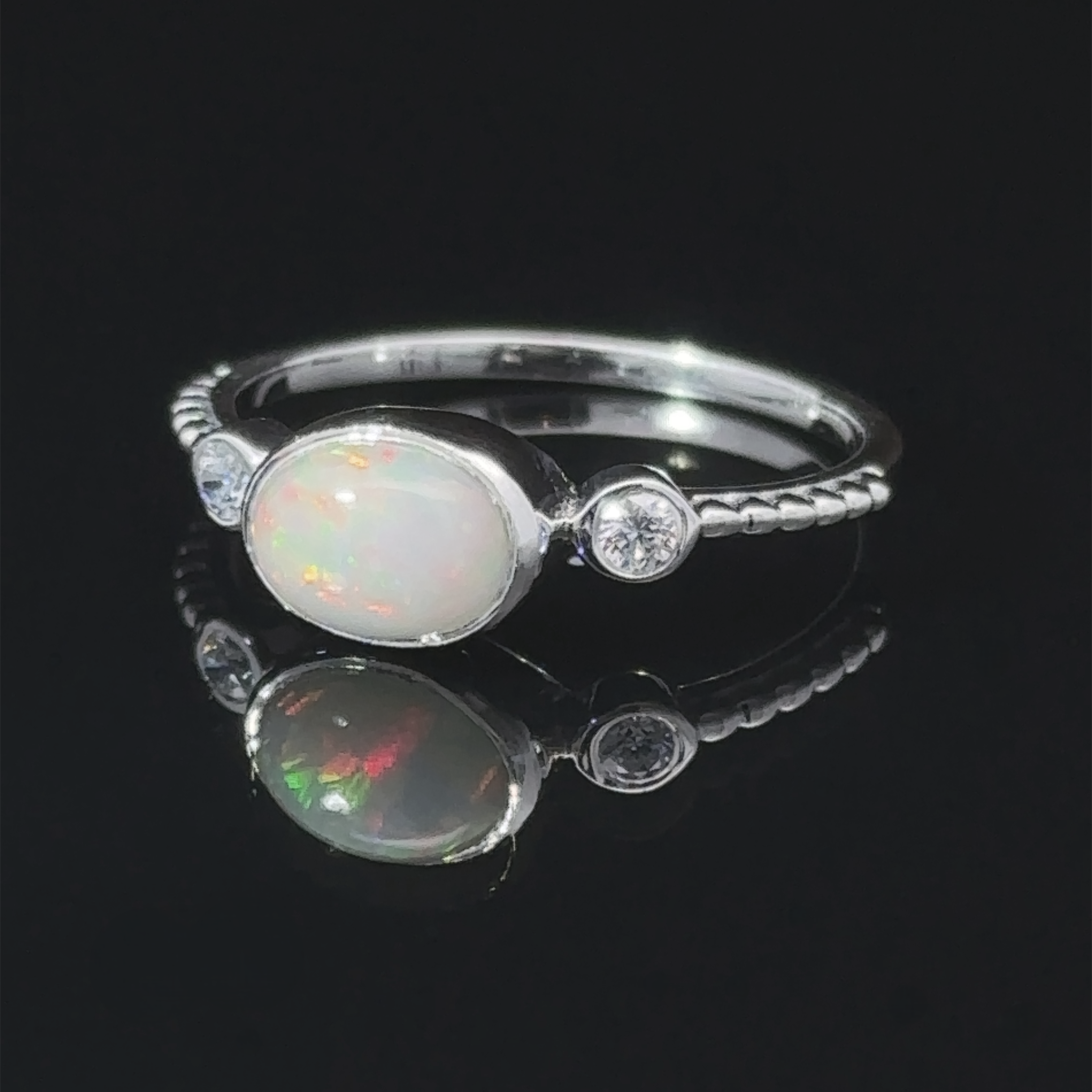 Horizontal Oval Gemstone Ring with Beaded Band with an opal center stone and small diamonds, displayed on a reflective surface.