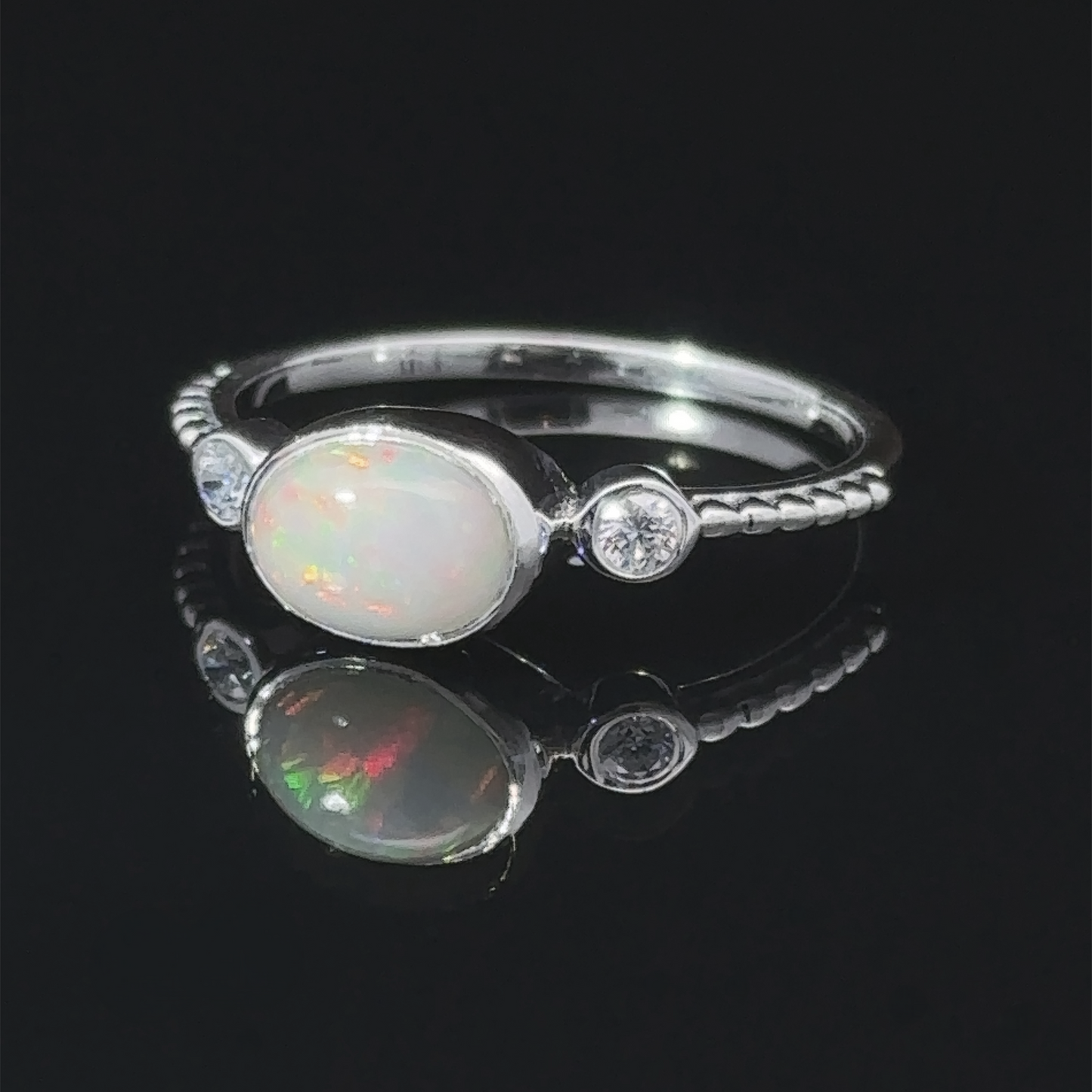 
                  
                    Horizontal Oval Gemstone Ring with Beaded Band with an opal center stone and small diamonds, displayed on a reflective surface.
                  
                