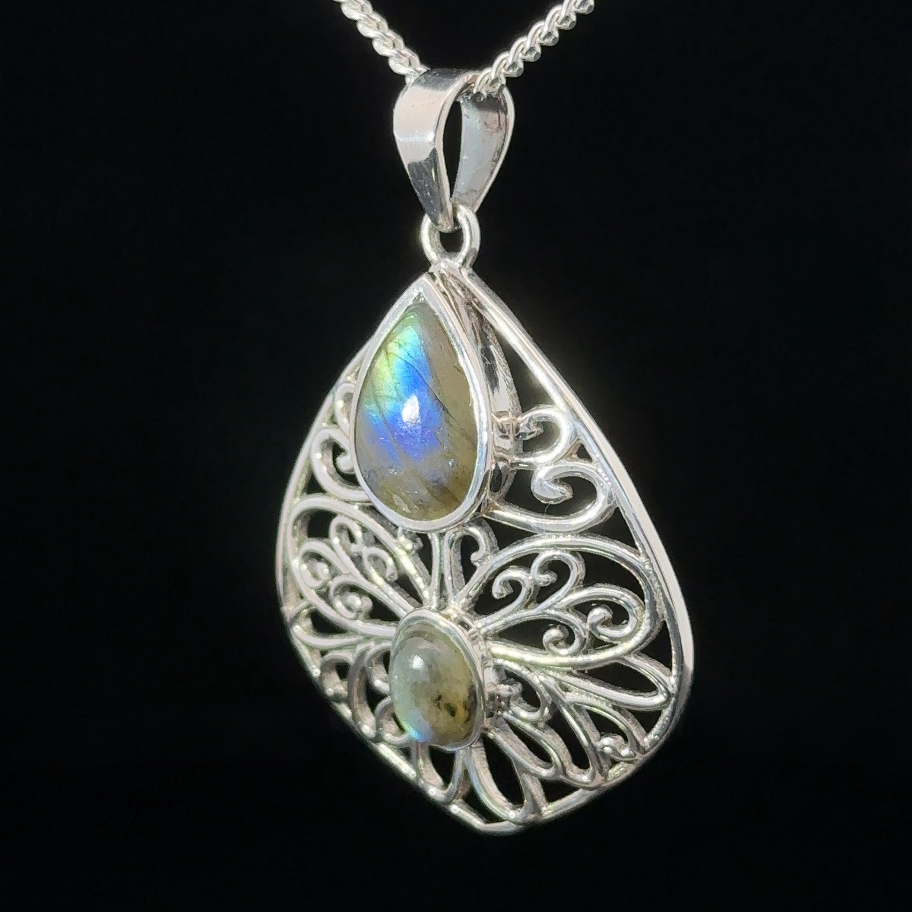 A Teardrop Filigree Gemstone Pendants with an intricate design featuring two labradorite stones, one teardrop-shaped and one oval-shaped, hangs from a silver chain against a black background. An amethyst accent adds a touch of mystique.