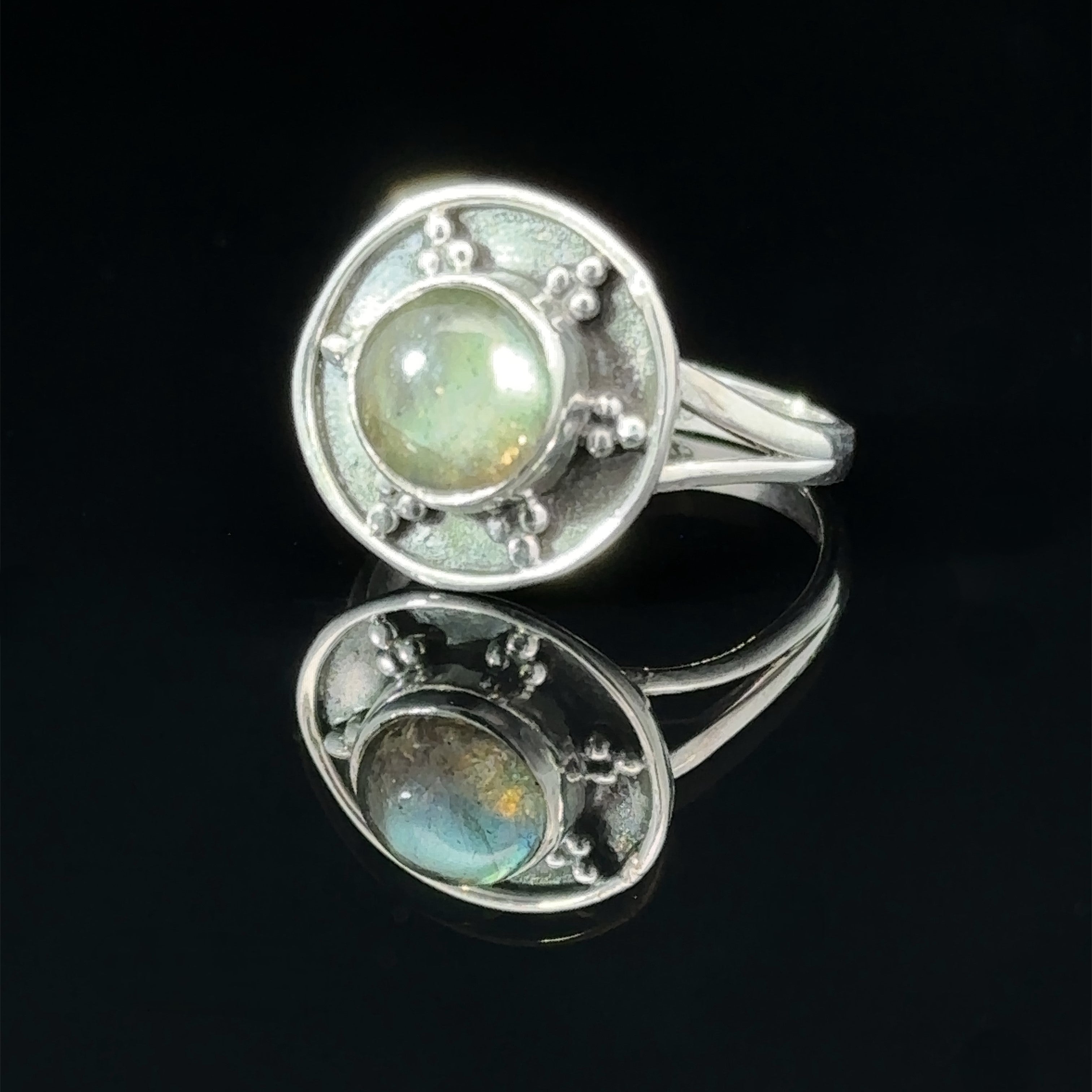 A Gemstone Ring With Unique Oxidized Design set in the center, featuring small circular details around the stone, is displayed against a black background with its reflection below.