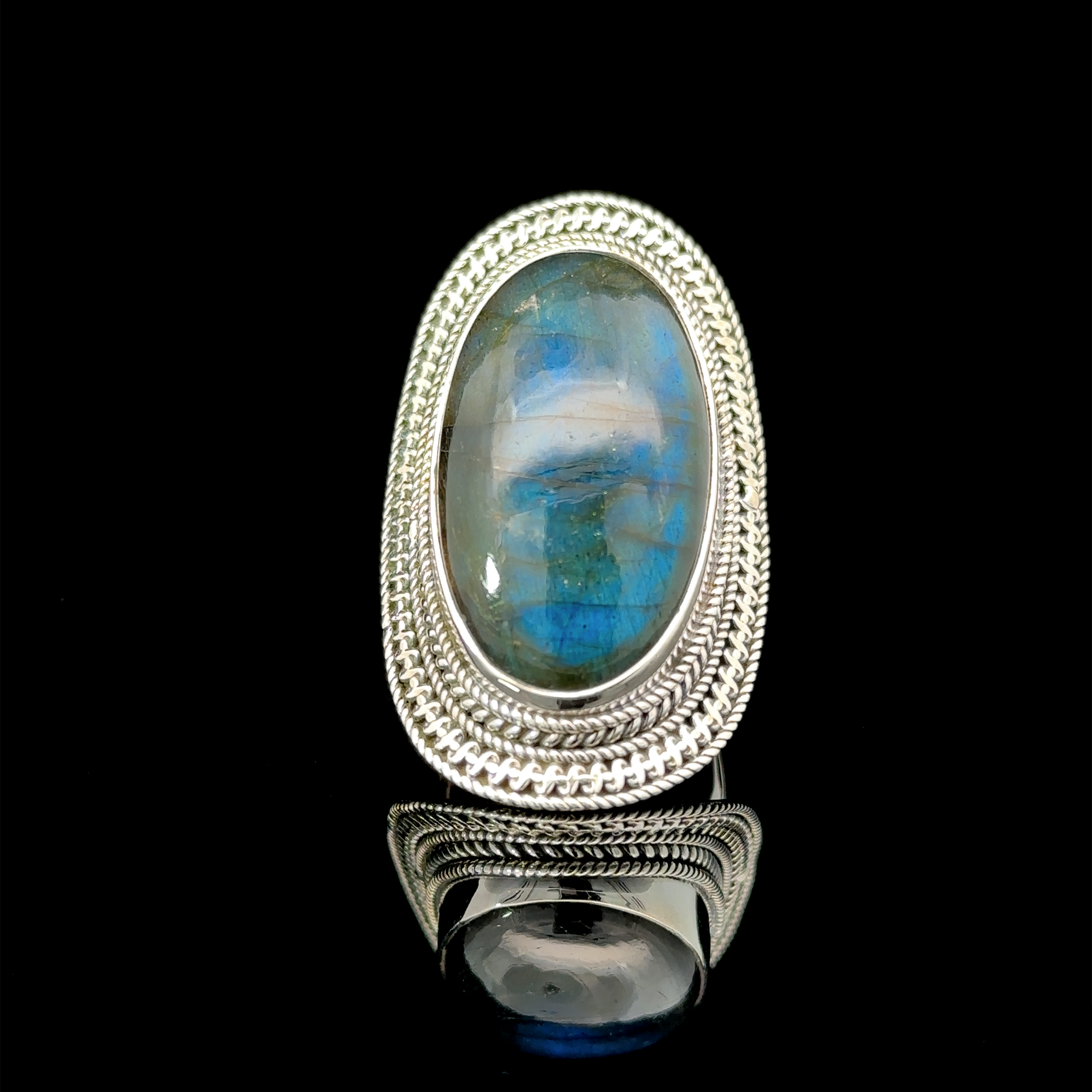 A Large Oval Shield Gemstone Ring with a large, oval-shaped blue-green gemstone, set in an intricately detailed band with a bohemian flair, displayed on a reflective black surface.