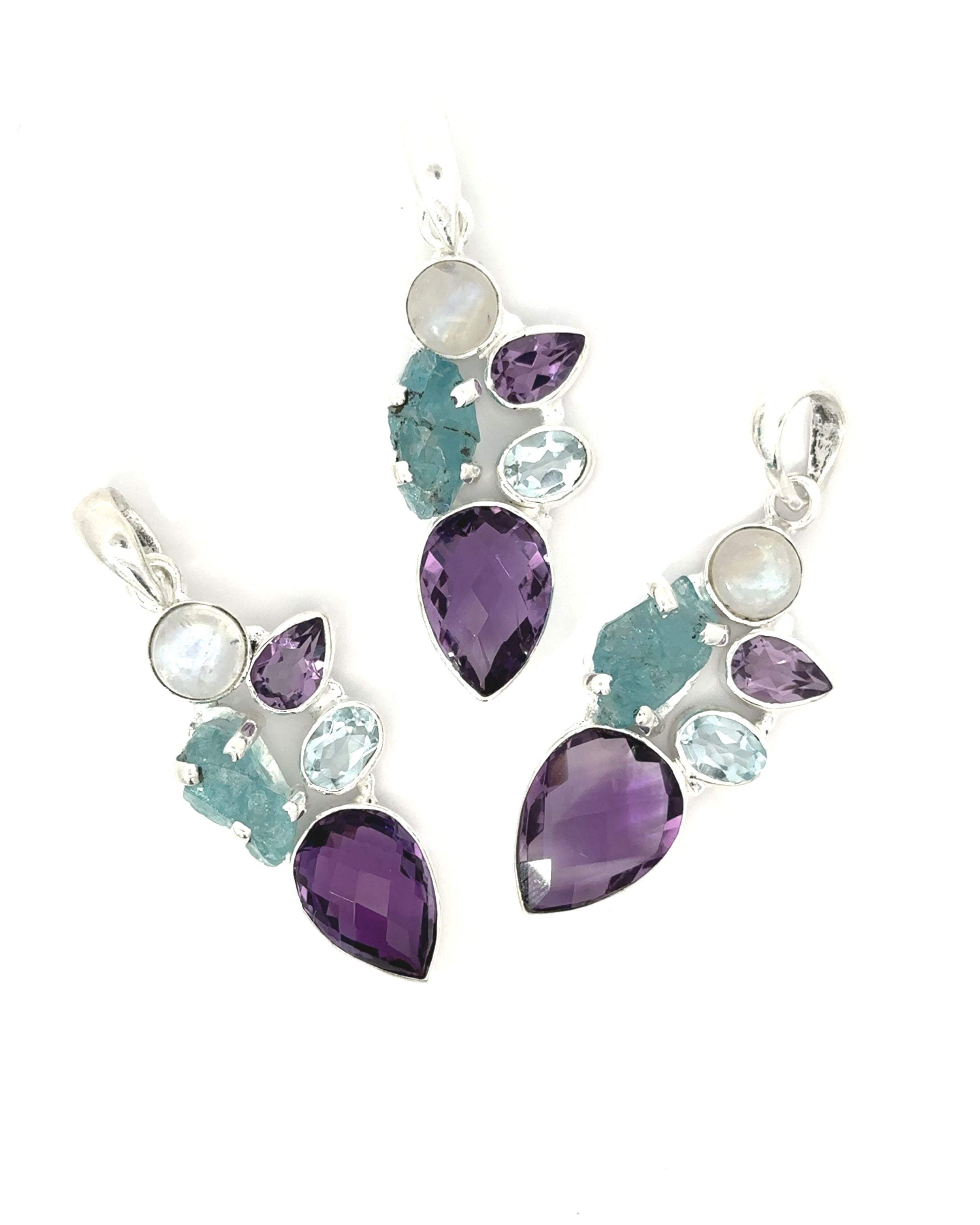 The Amethyst Multi Stone Pendant presents three silver pendants with an abstract design on a white background, embracing a bohemian jewelry style. It features a vibrant mix of purple, teal, and clear gemstones in teardrop and oval shapes, echoing the essence of an amethyst pendant.