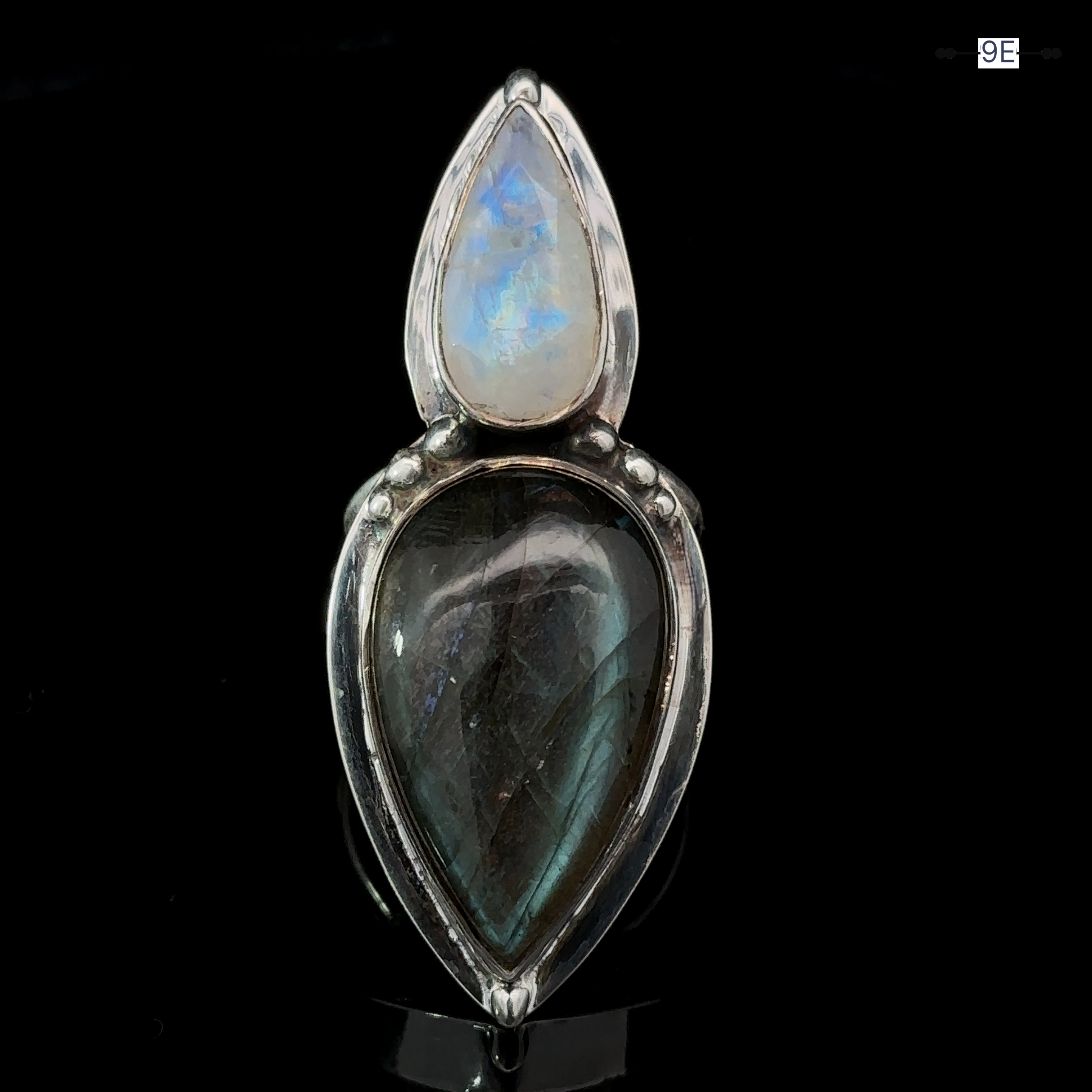 The Heavy Labradorite And Moonstone Ring is a silver statement piece featuring two tear-shaped gemstones; the upper stone is a moonstone with a translucent blue sheen, while the lower stone is labradorite with a darker greyish hue, set against a black background.