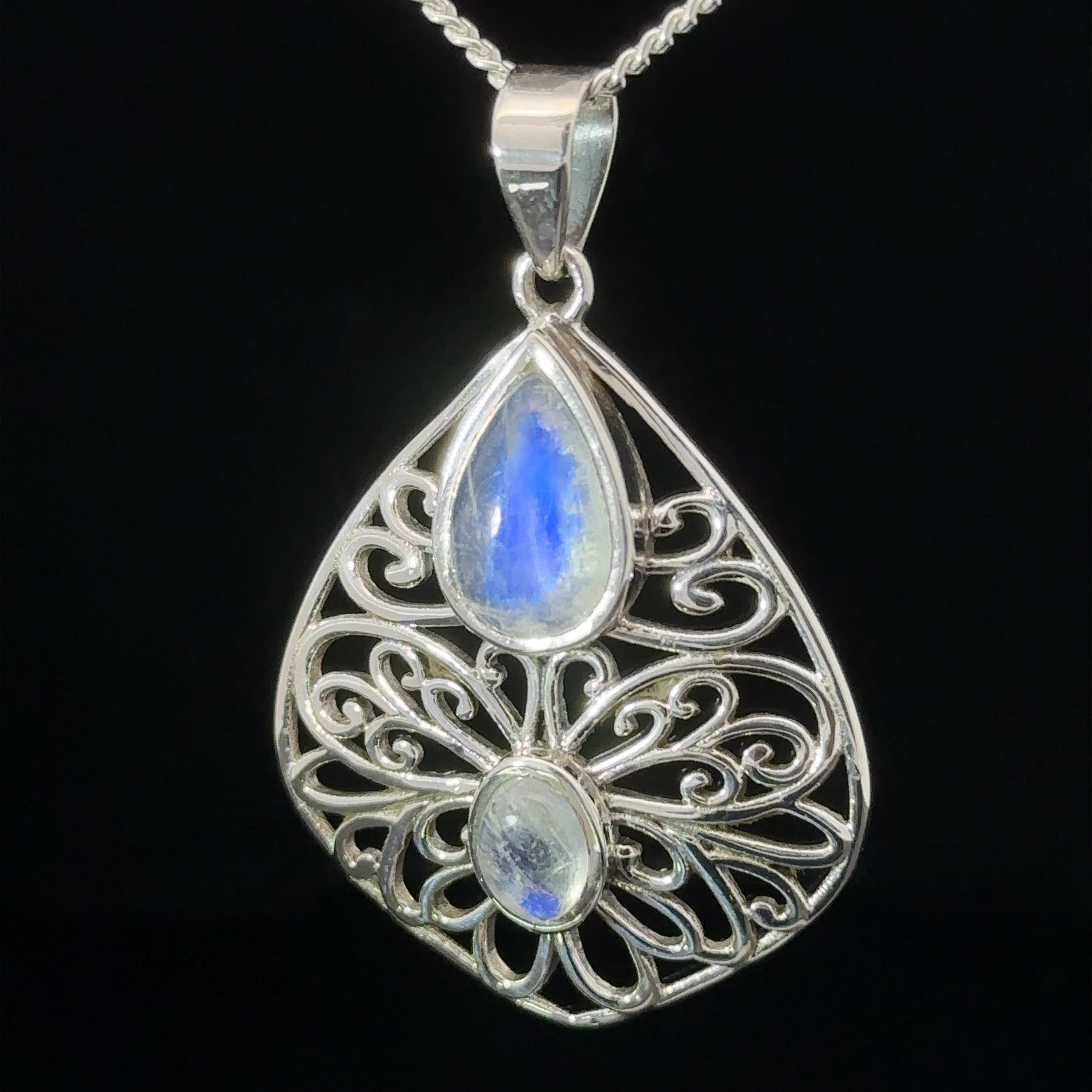 The **Teardrop Filigree Gemstone Pendants** with intricate filigree design featuring two blue gemstones, one teardrop-shaped at the top and an oval one below, hanging from a silver chain against a black background.