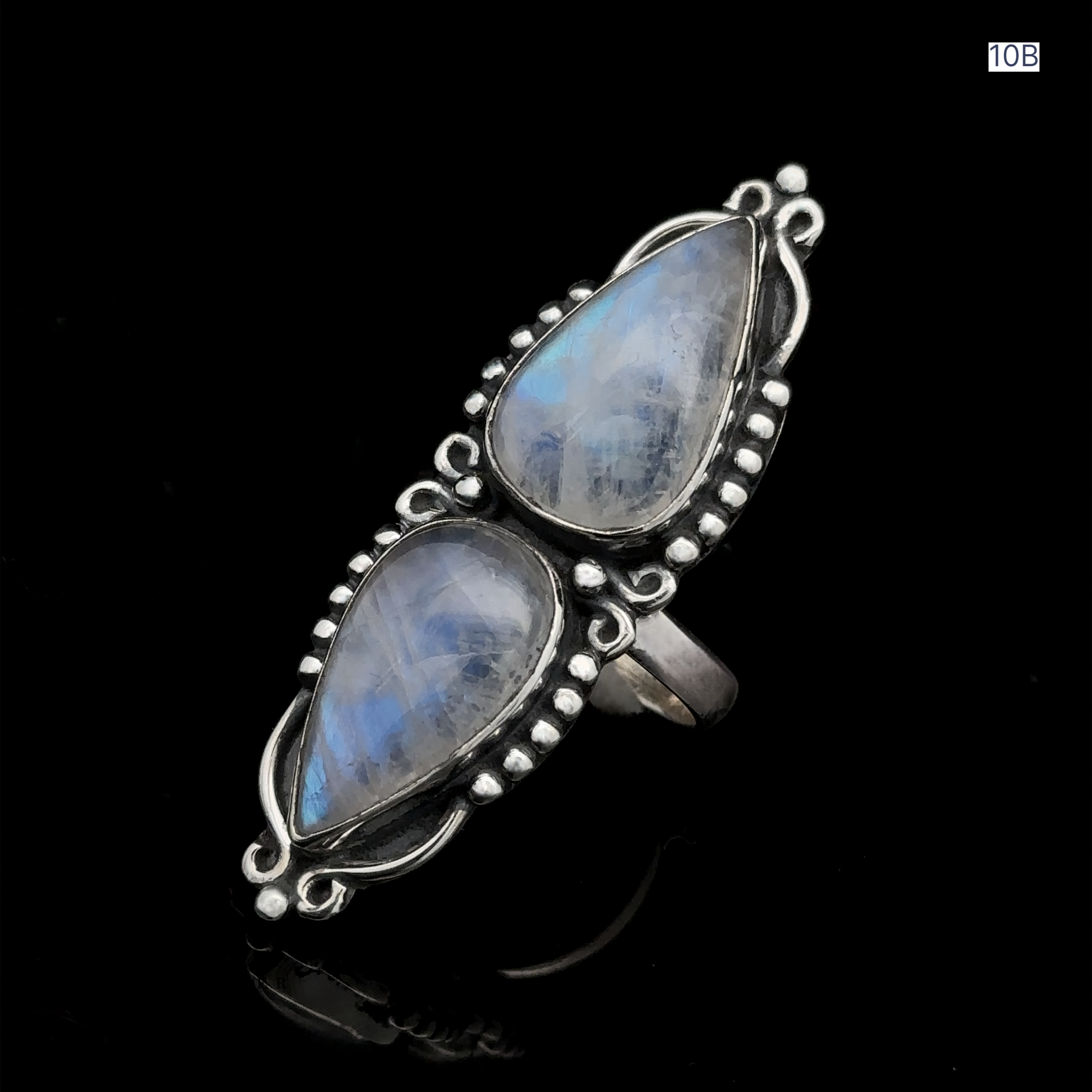 The Long Double Teardrop Moonstone Ring showcases two vertically set, pear-shaped translucent moonstones, enhanced by detailed metalwork and small metallic beads on a black background.