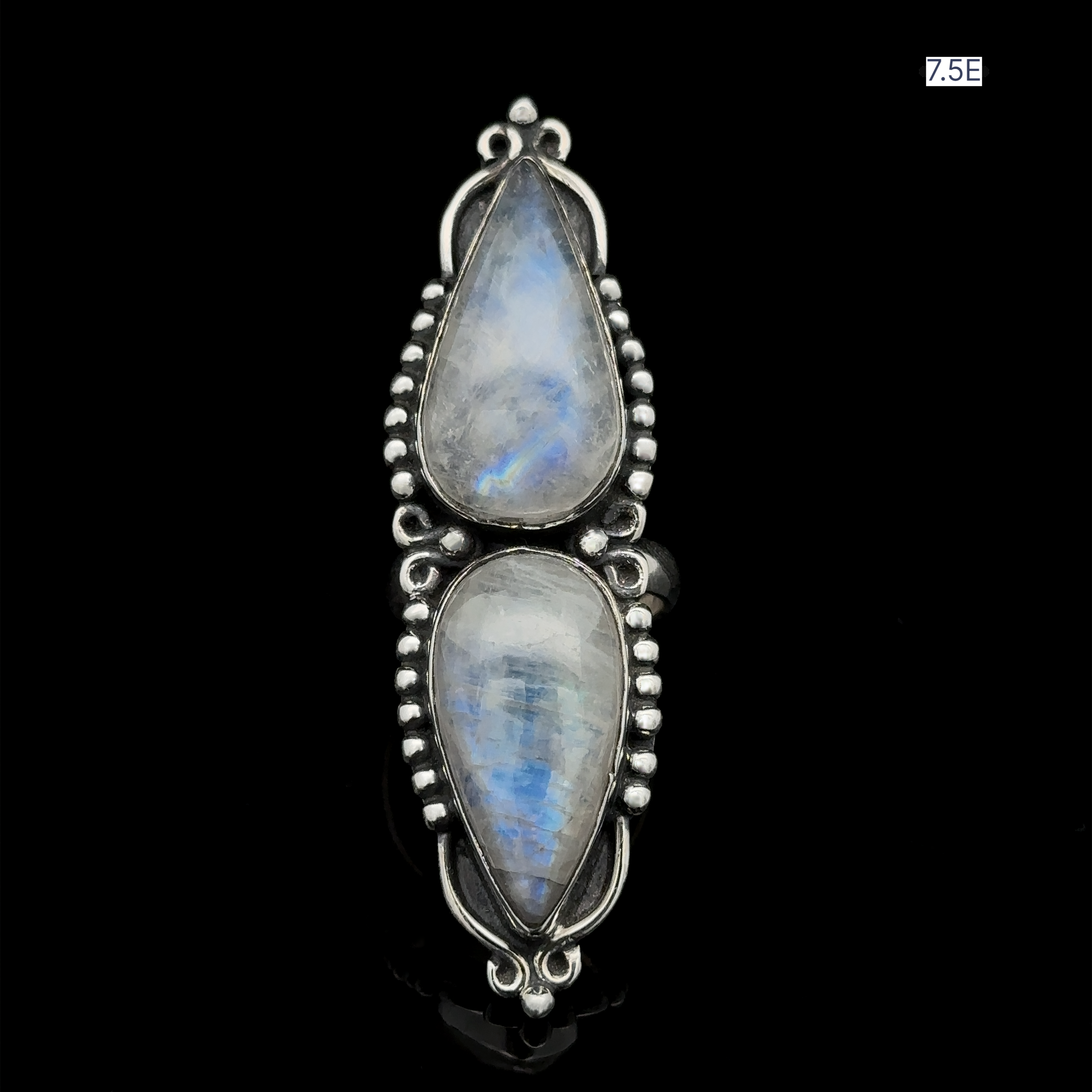 The Long Double Teardrop Moonstone Ring features two vertically set pear-shaped moonstones, enveloped by elaborate metalwork. The label "7.5E" is distinct in the top right corner, highlighting its elegance and expert craftsmanship, making it an ideal accessory for any occasion.