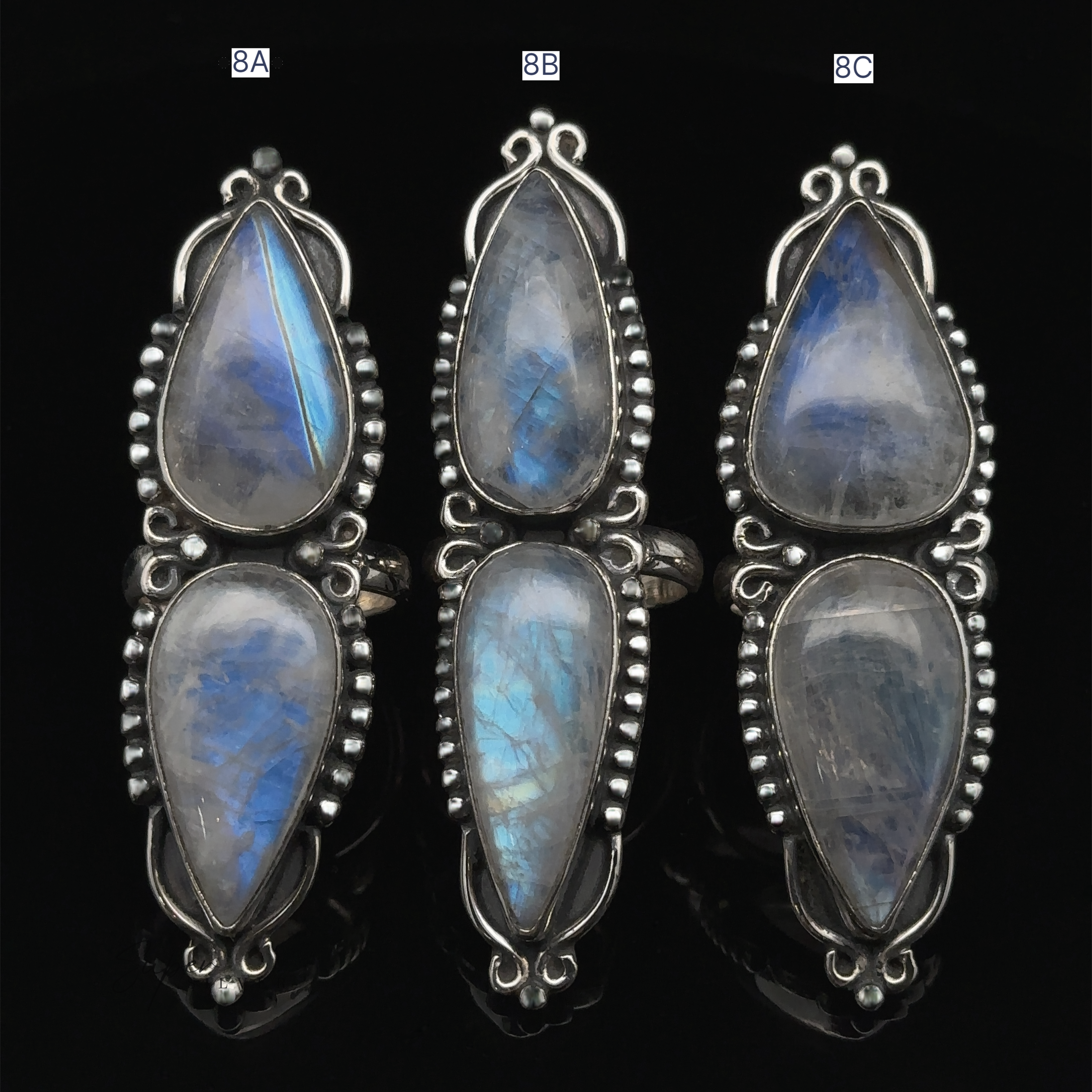 Three Long Double Teardrop Moonstone Rings, each with two oval rainbow moonstones, are arranged vertically against a black background. The rings are labeled 8A, 8B, and 8C.