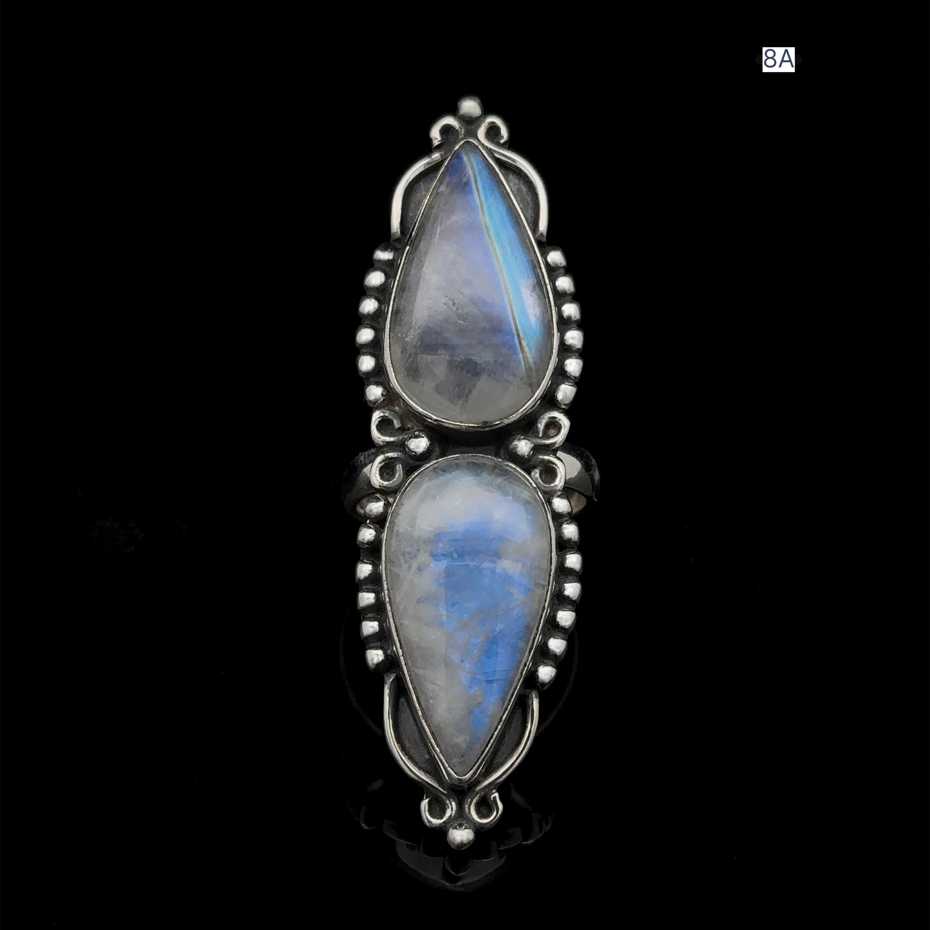 The Long Double Teardrop Moonstone Ring, crafted from sterling silver, showcases two tear-shaped moonstones with a blue tint, each encased in an ornate metal frame and set against a black background.