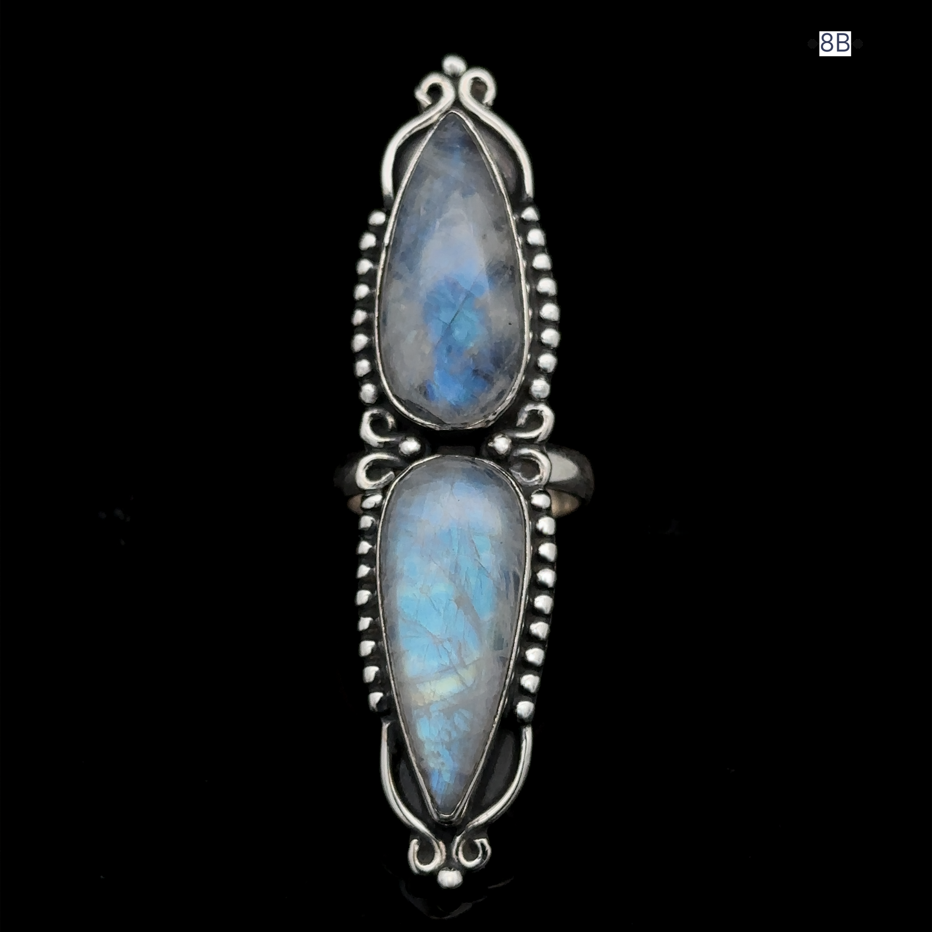 The Long Double Teardrop Moonstone Ring, crafted from sterling silver, showcases two vertically aligned pear-shaped moonstones set in an ornate bezel adorned with delicate silver accents against a black background—this statement piece demands attention.