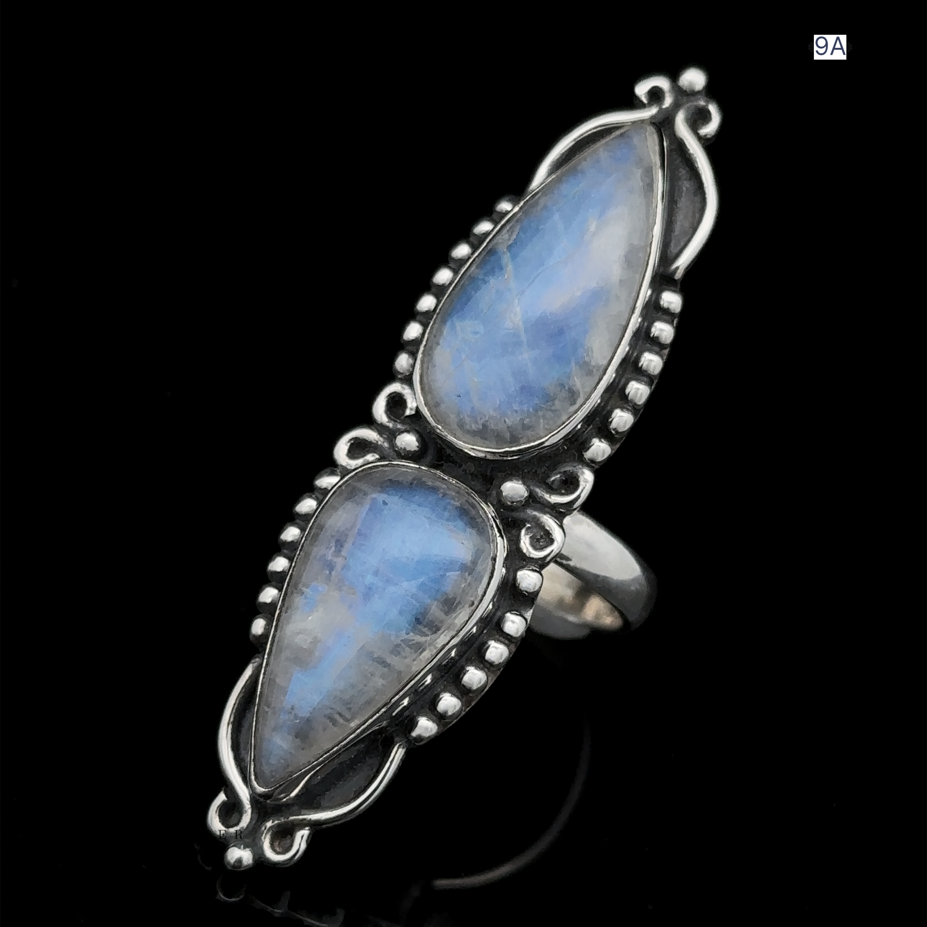 The Long Double Teardrop Moonstone Ring is a sterling silver statement piece showcasing two vertically set, pear-shaped blue moonstone gemstones, adorned with intricate metalwork around each stone and displayed against a black background.