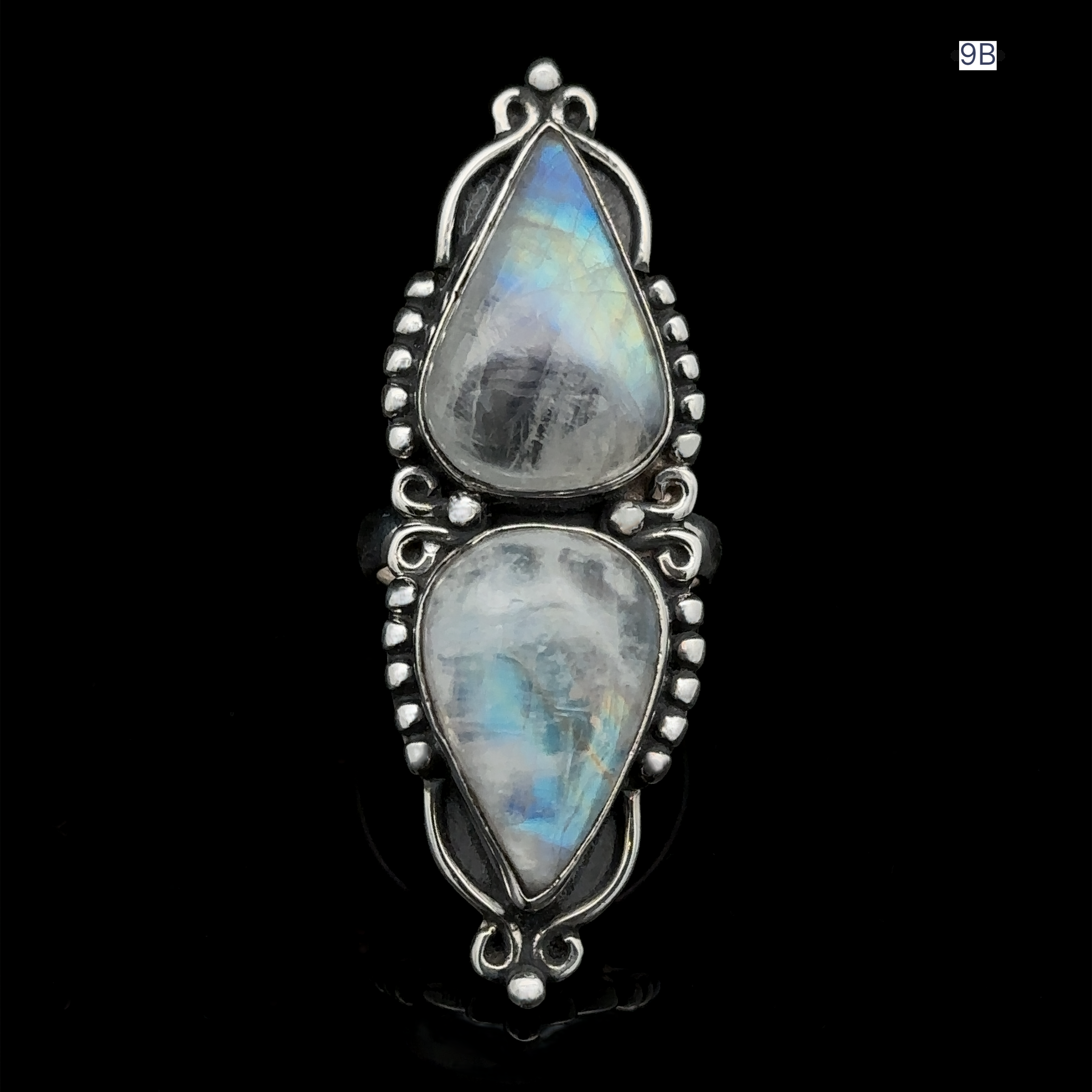 The Long Double Teardrop Moonstone Ring showcases two vertically-set, semi-transparent pear-shaped moonstones, framed by intricate sterling silver metalwork against a black background. This eye-catching and elegant statement piece is sure to impress.