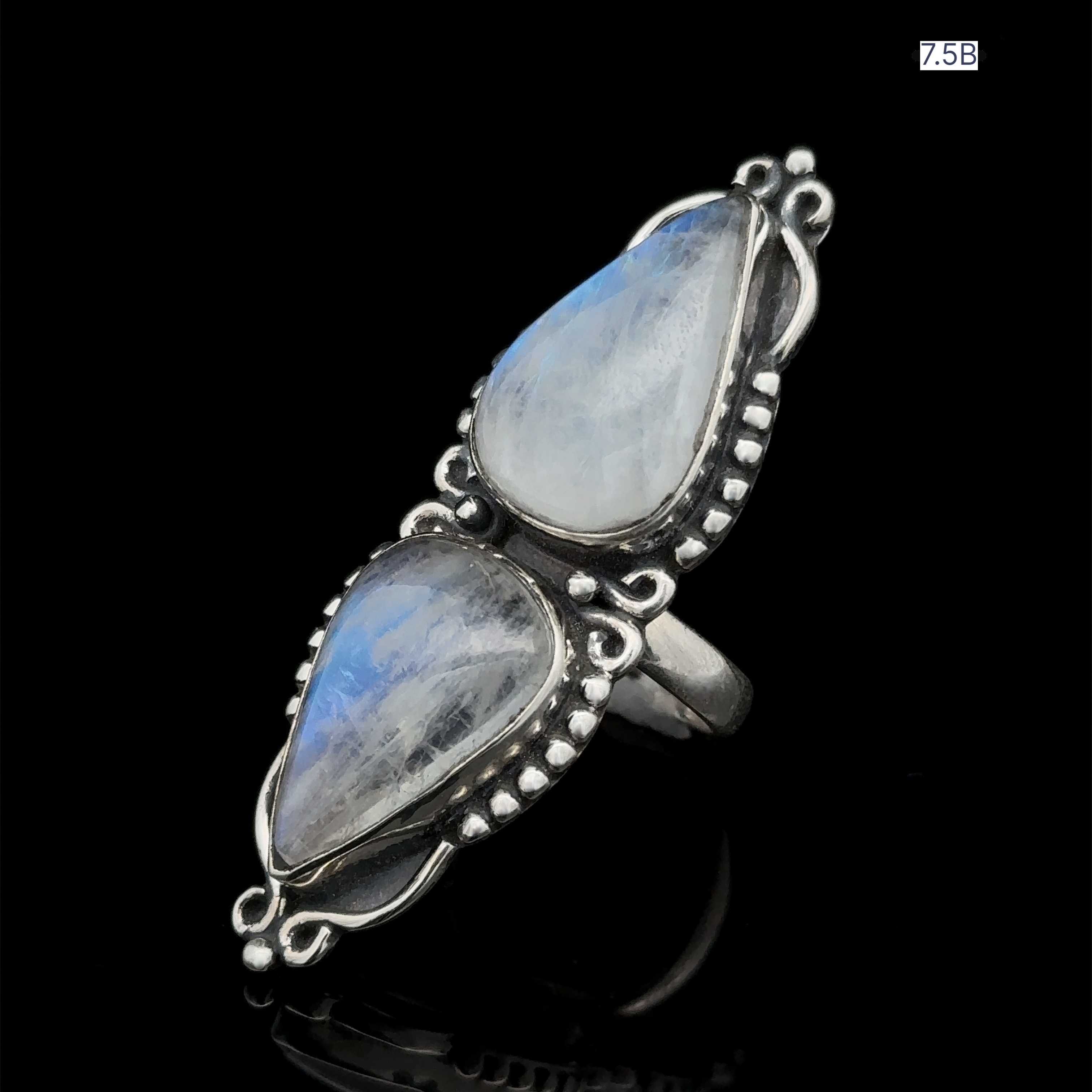 The Long Double Teardrop Moonstone Ring is a striking statement piece featuring a silver double teardrop moonstone with ornate detailing, set against a black background. It is crafted from sterling silver and is available in size 7.5.