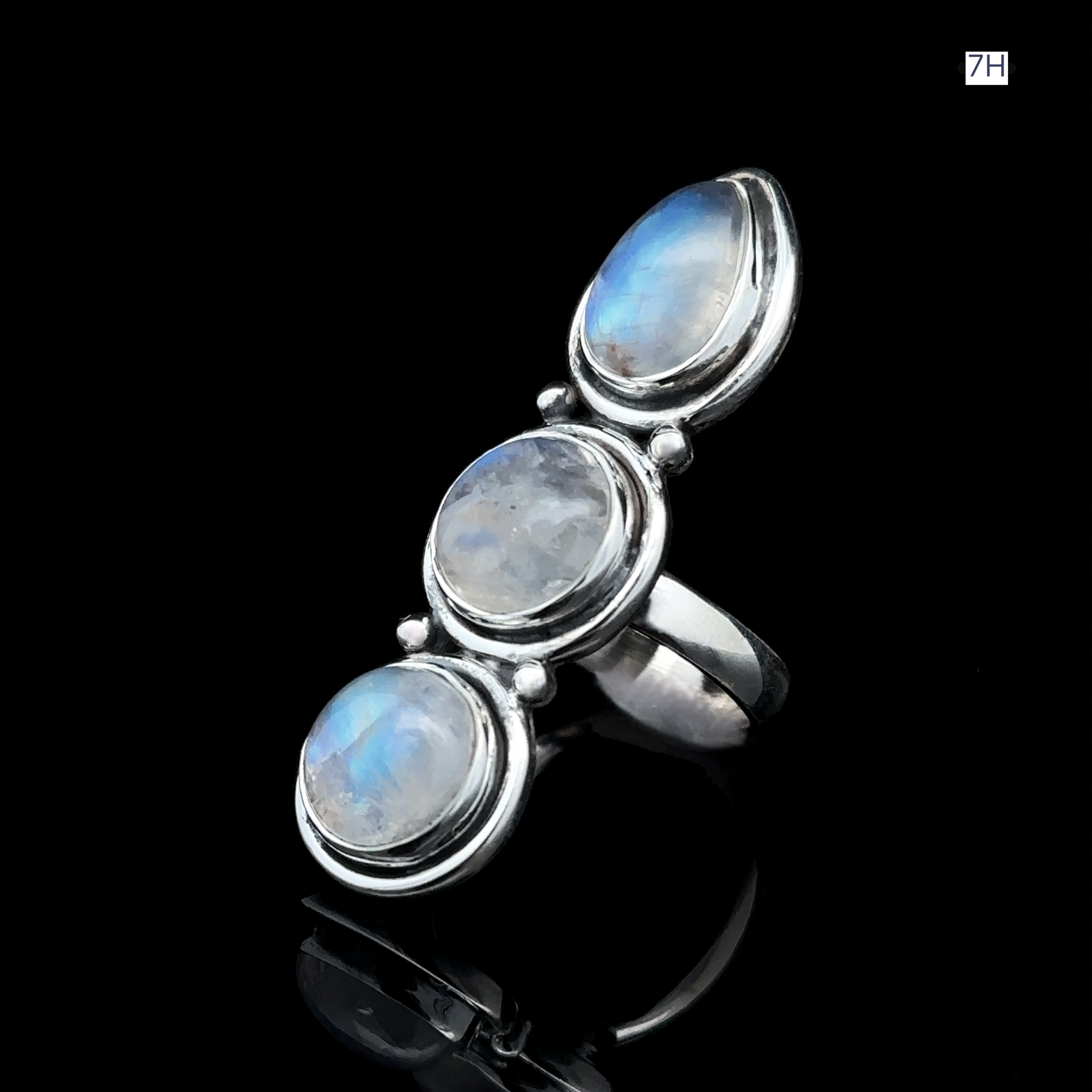 The Triple Moonstone Ring, crafted from sterling silver and featuring three vertically set oval moonstones, radiates goddess energy as it gleams on a black reflective surface.