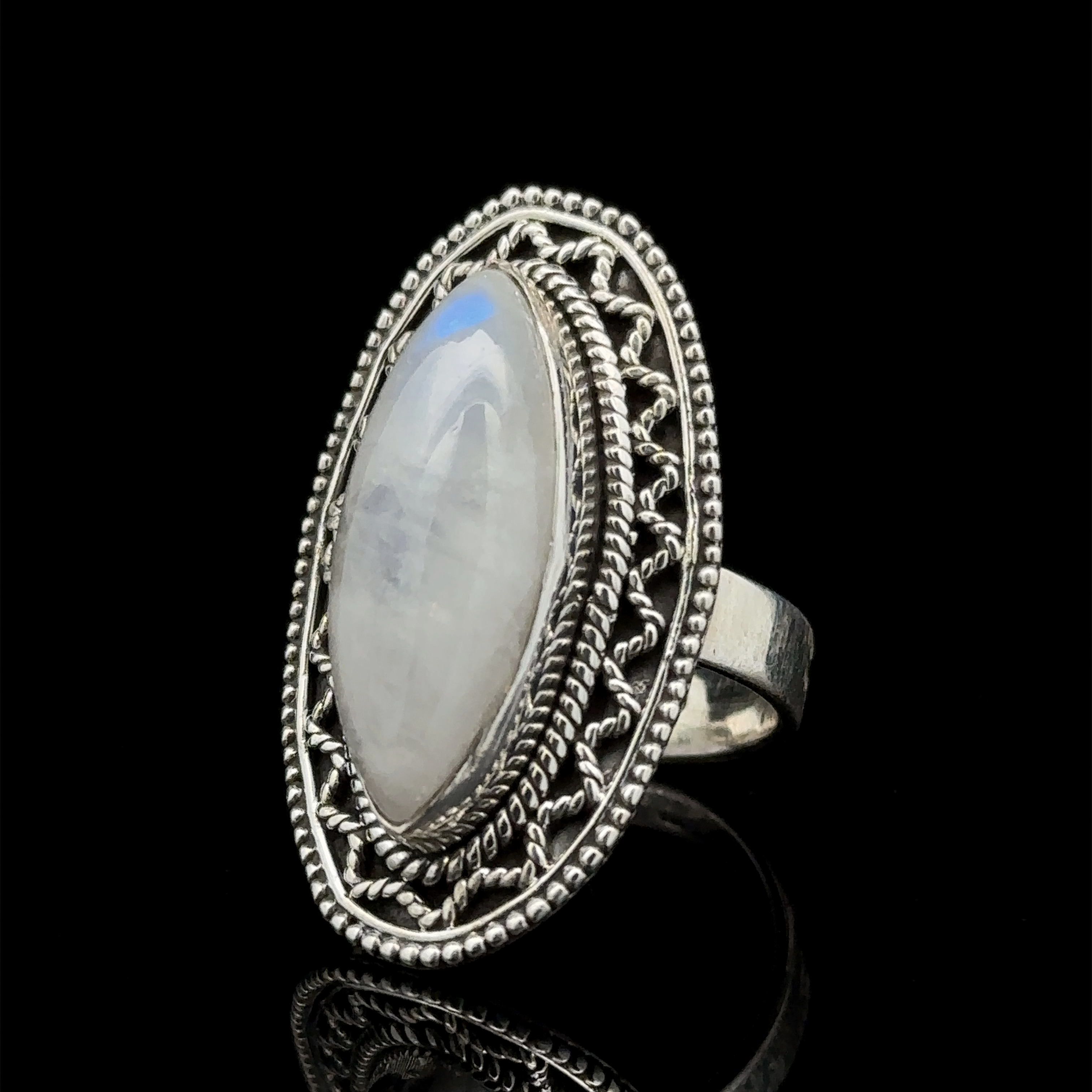 An oval-shaped moonstone set in an intricately designed sterling silver ring with a detailed band and filigree pattern, displayed against a black background, offers a subtle Bohemian twist of the Marquise Shaped Gemstone Ring With Vintage Shield Border.