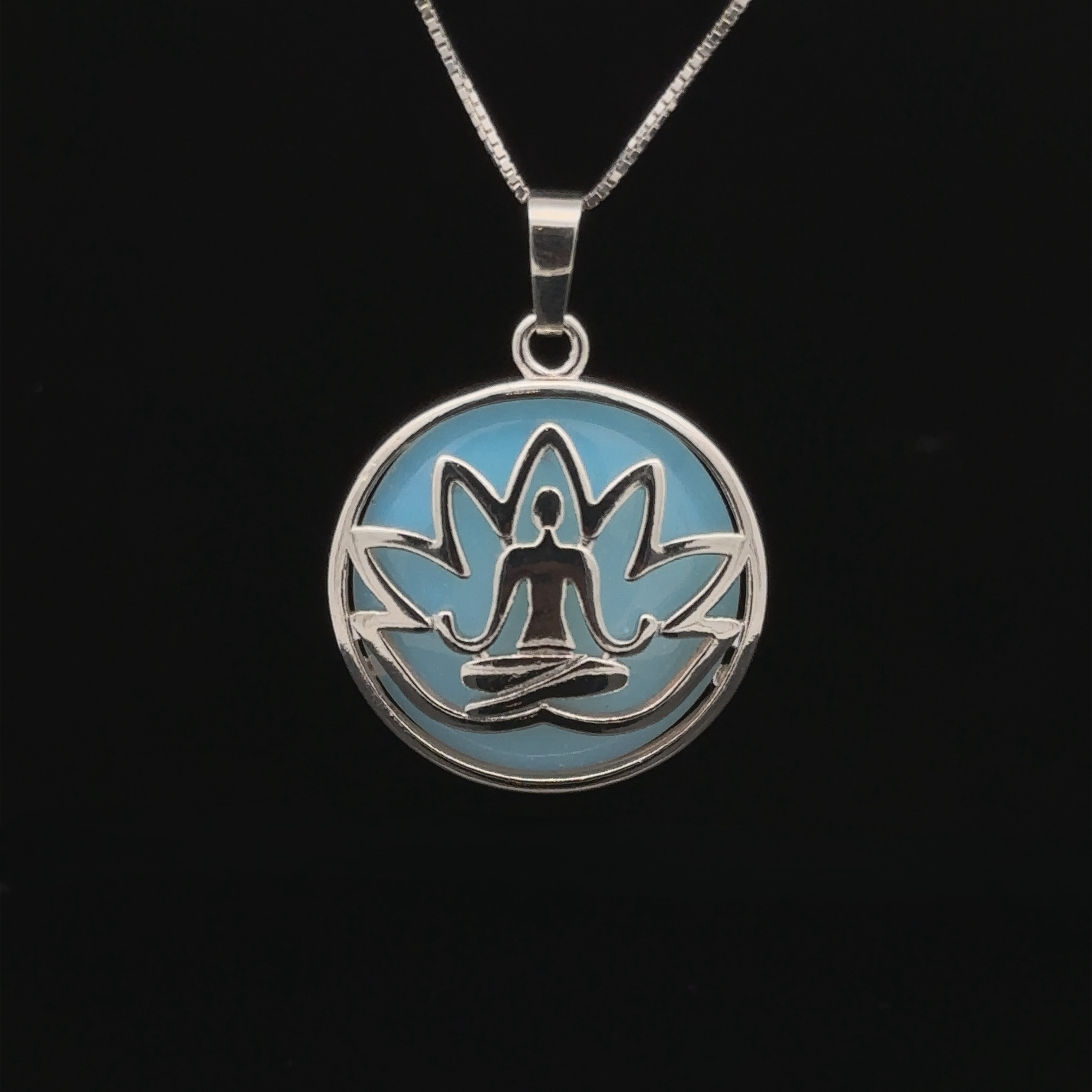 A Silver Plated Lotus Meditation Pendant with Gemstone features a lotus design with a seated meditation figure at its center, set against a light blue background. The pendant hangs on a thin silver chain. Black background.