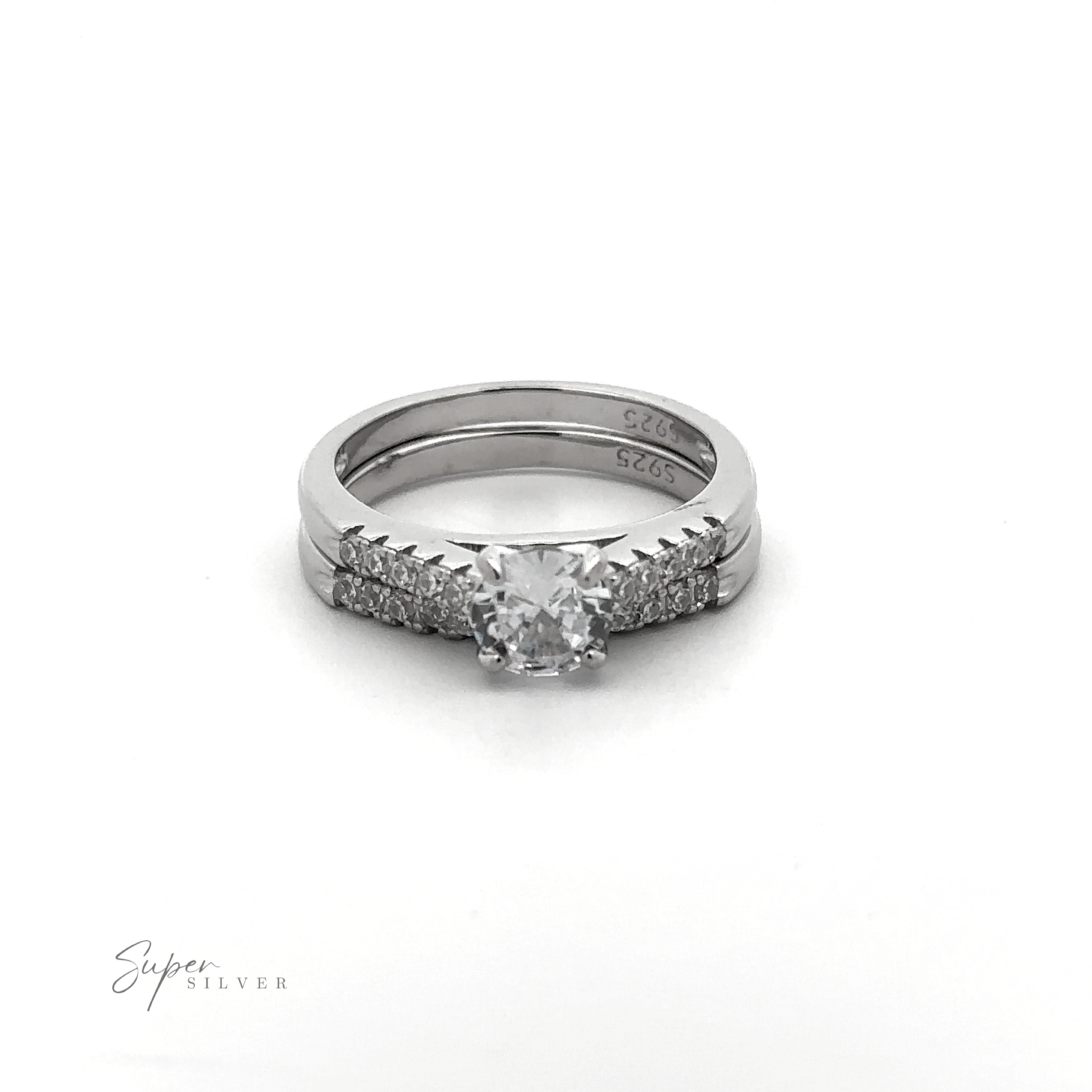 The Round Solitaire Wedding Band Set features a central diamond with small diamonds adorning the sterling silver band, all displayed atop a plain white background. The rhodium finish adds an extra touch of elegance to this timeless piece.