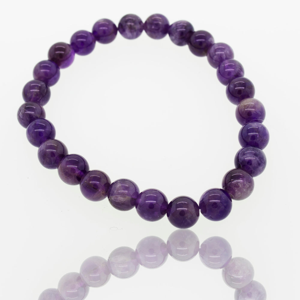 
                  
                    A 4mm Beaded Stone Bracelet made of glossy purple amethyst beads, known as a healing stone bracelet, arranged in a circular shape and displayed on a reflective white surface.
                  
                